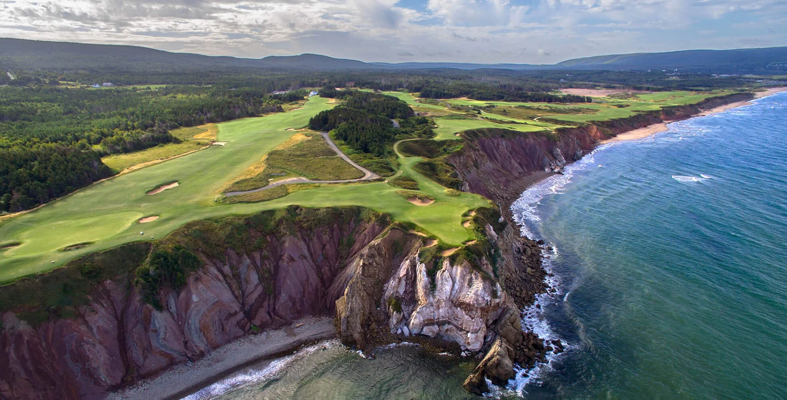 Paysage côtier avec un terrain de golf perché sur des falaises rocheuses, vue sur la mer avec vagues, zones de végétation, et montagnes en arrière-plan.