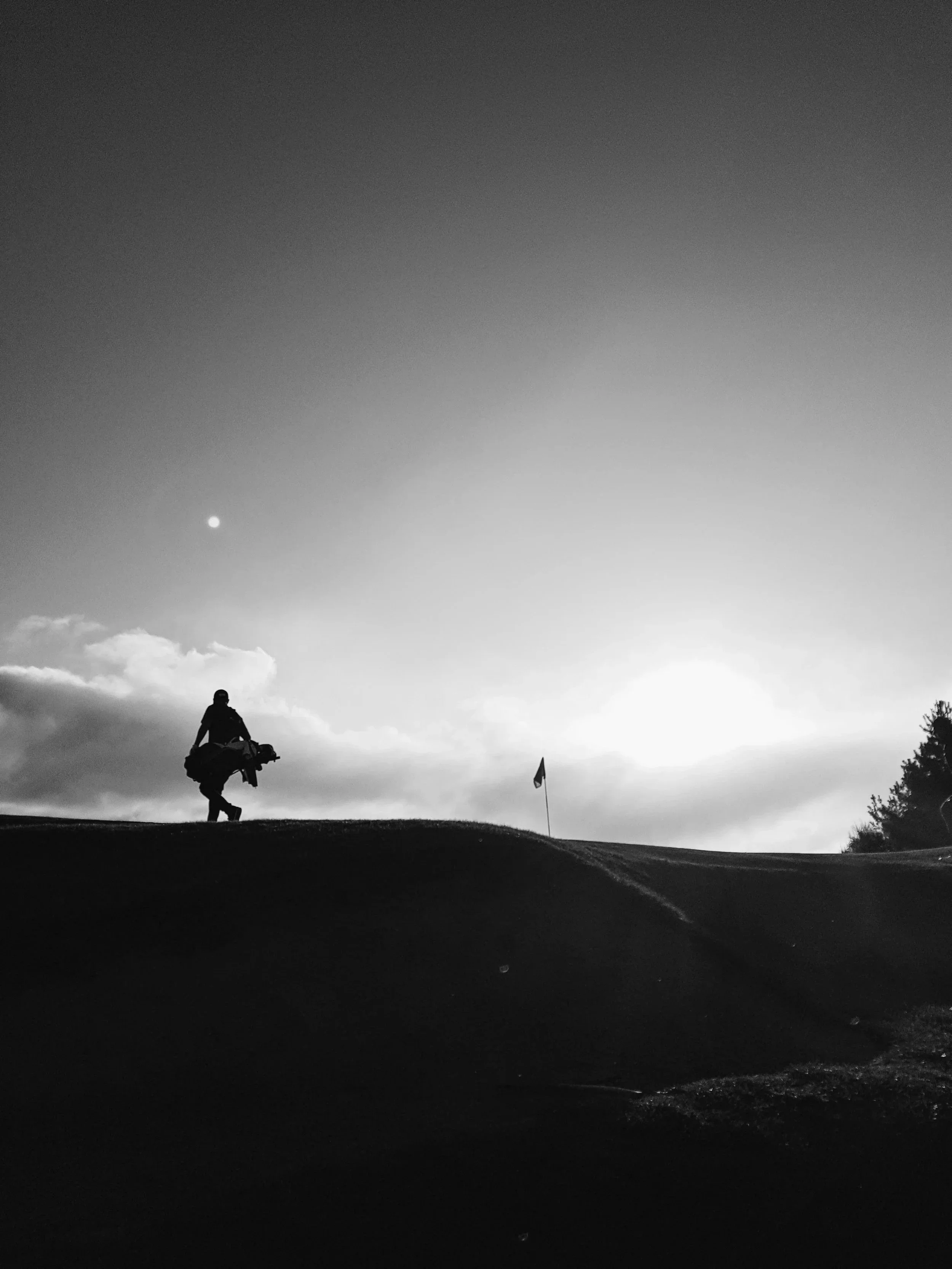 Silhouette d'un golfeur marchant avec un sac de golf sur un terrain de golf au coucher du soleil, avec un drapeau de trou visible et un ciel nuageux en arrière-plan.