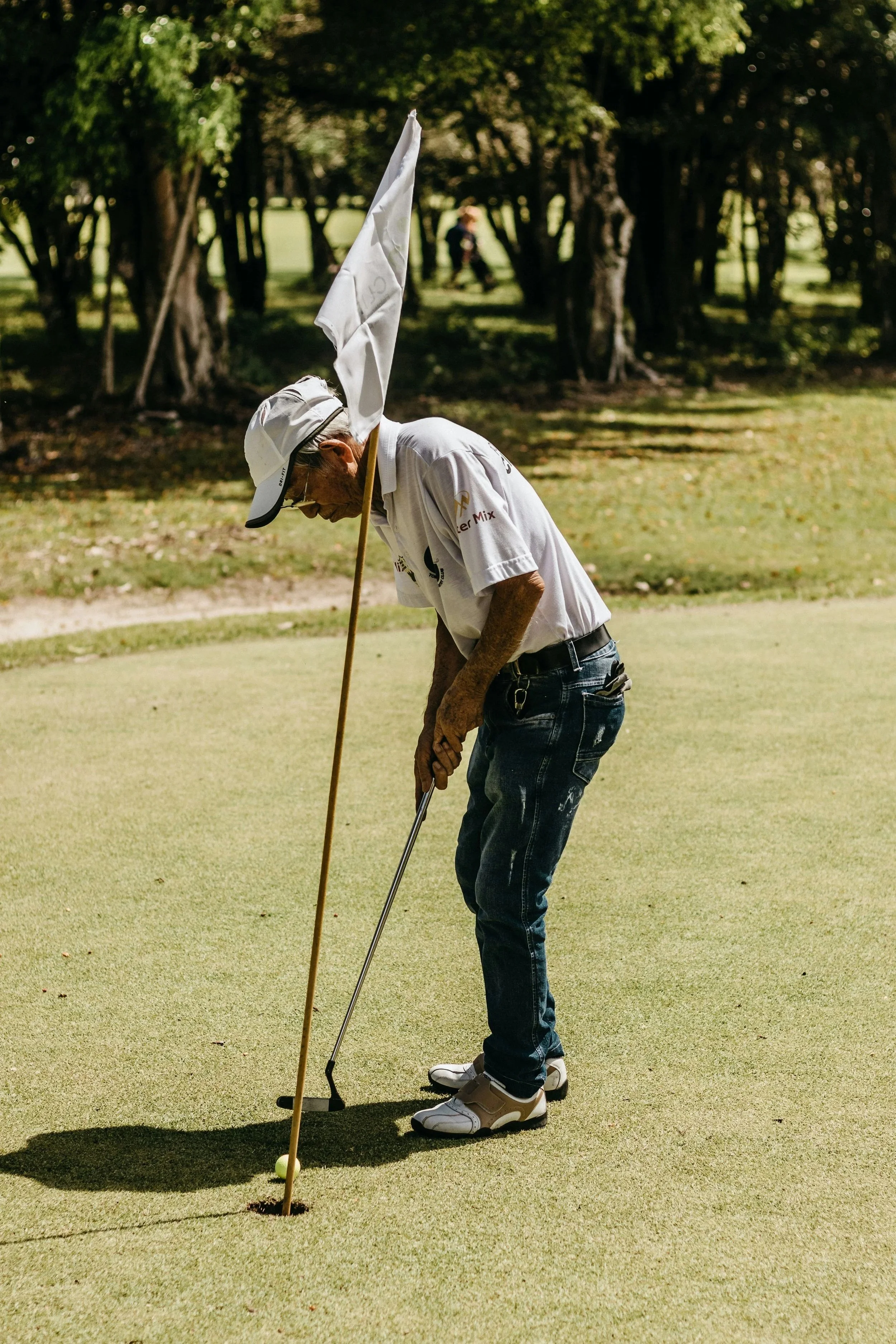 A senior man playing golf on a lush green course, preparing to putt the ball into the hole, with trees in the background.