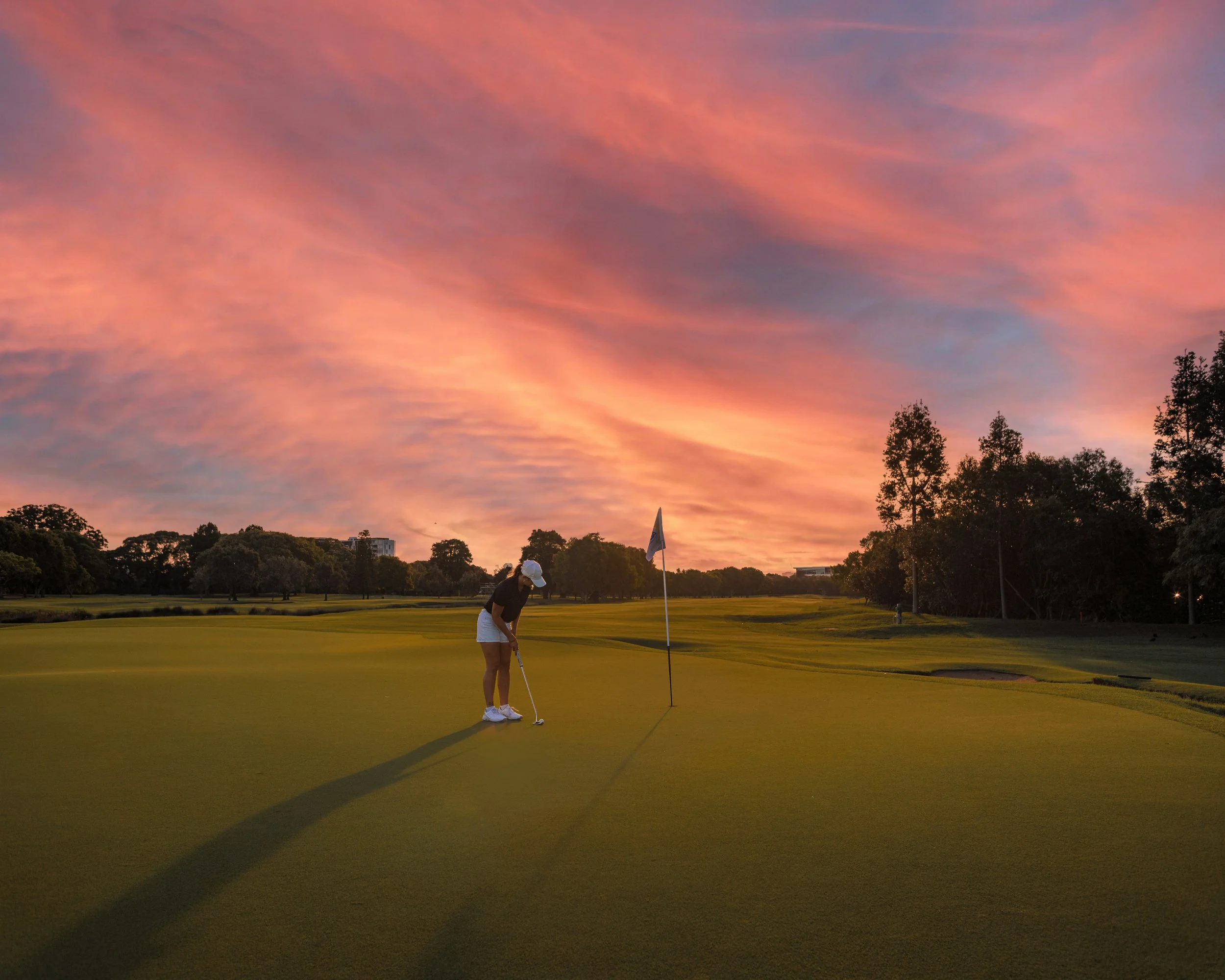Une joueuse de golf prépare son coup sur le green lors d'un coucher de soleil aux couleurs roses et orange, avec des arbres en arrière-plan.