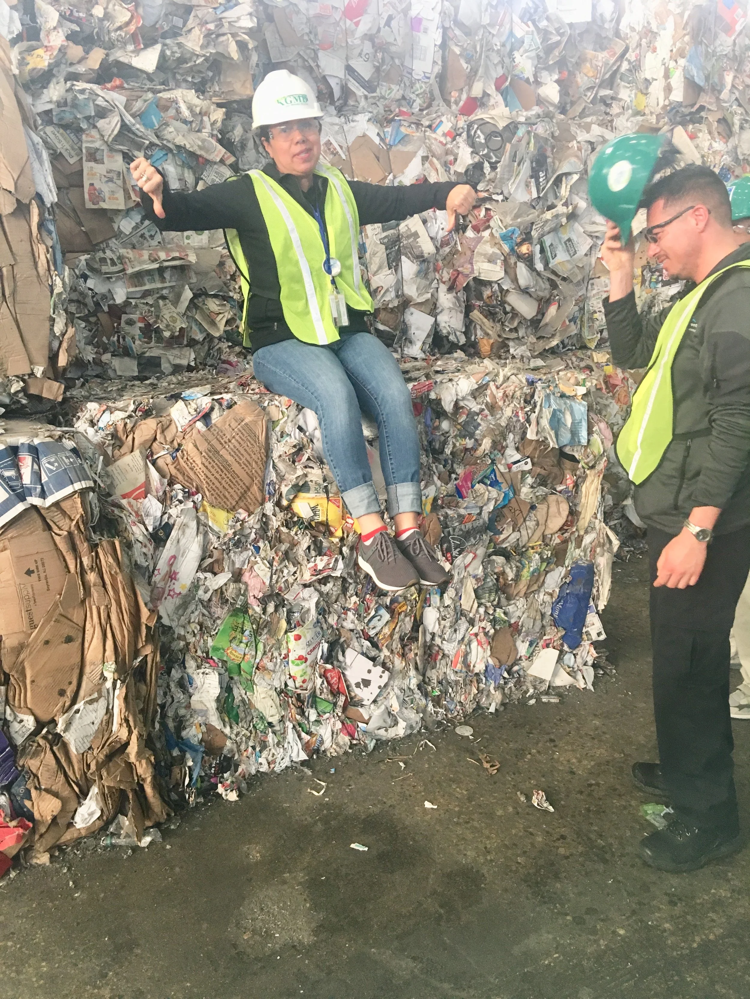 Two people wearing yellow safety vests and hard hats inside a large recycling or waste management facility. One person is sitting on a pile of crumpled paper and cardboard, holding the hard hat in her hand. The other person is standing nearby, also wearing safety gear, with a smile on his face.