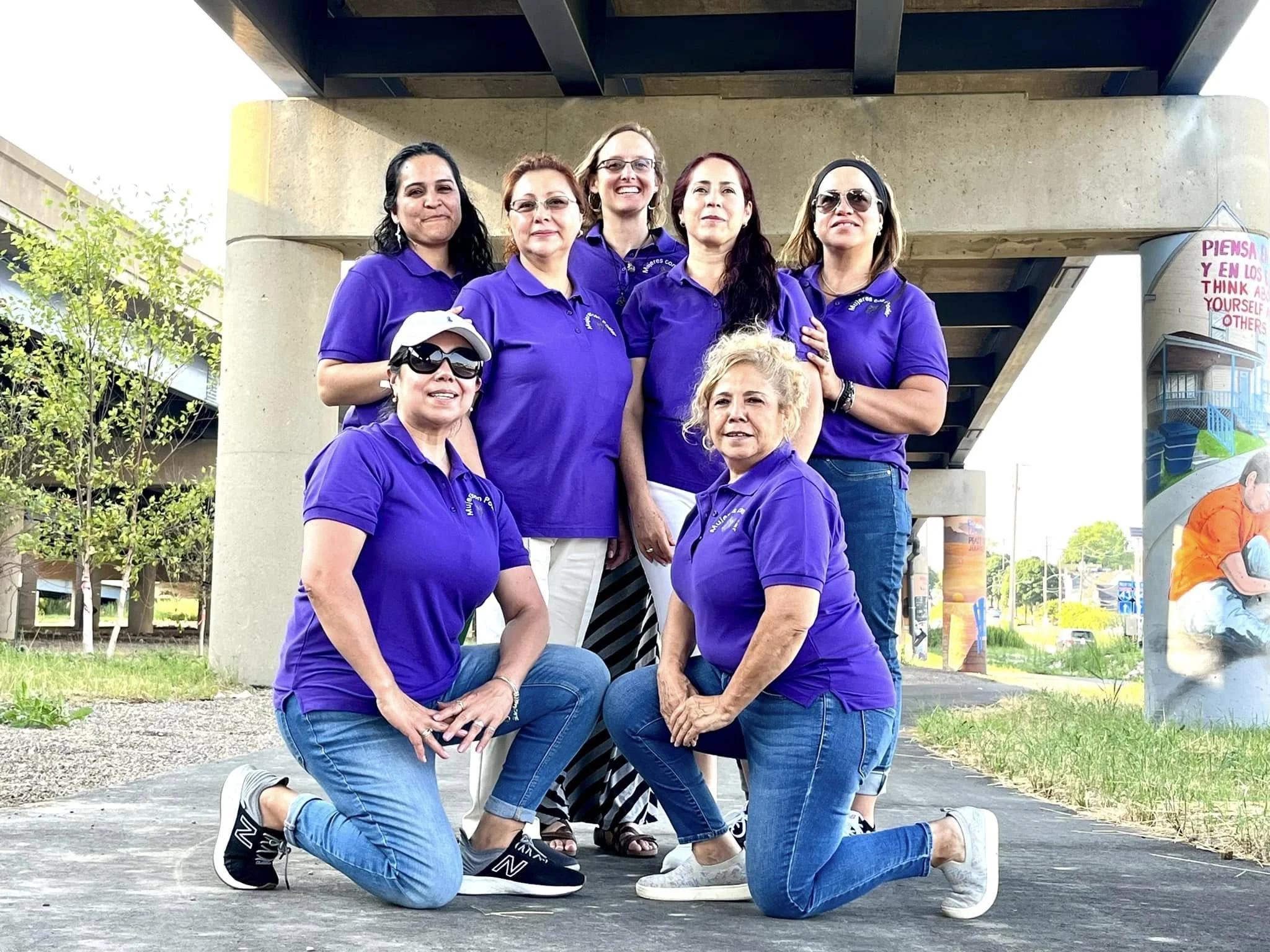A group of eight women wearing matching purple shirts, posing outdoors under a bridge. Some are standing, while others are kneeling or crouching, smiling at the camera. In the background, graffiti art and trees are visible.