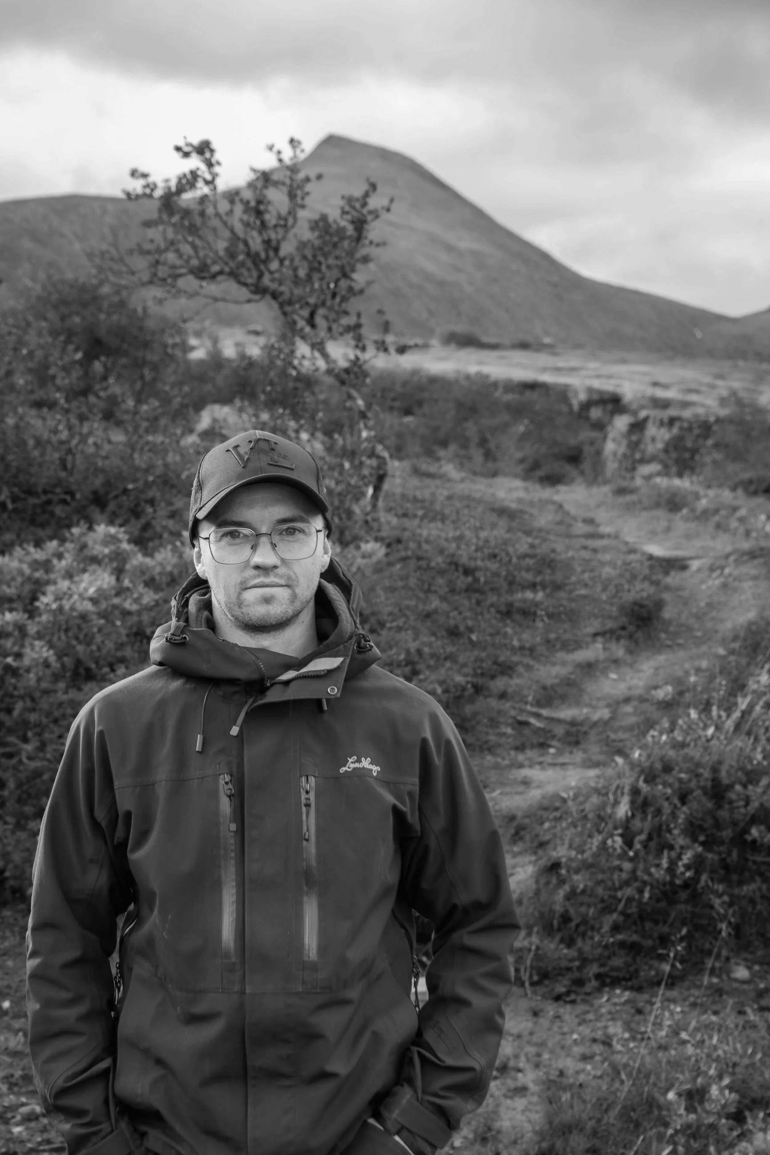 En man i jacka, keps och glasögon står framför en landskap med en stig och berg i bakgrunden, svartvitt.