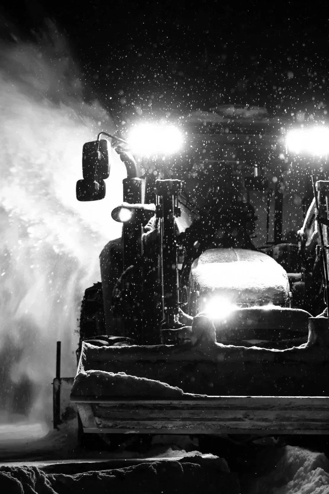 En snötäckt traktor med starka ljus, som arbetar i mörker under snöväder.