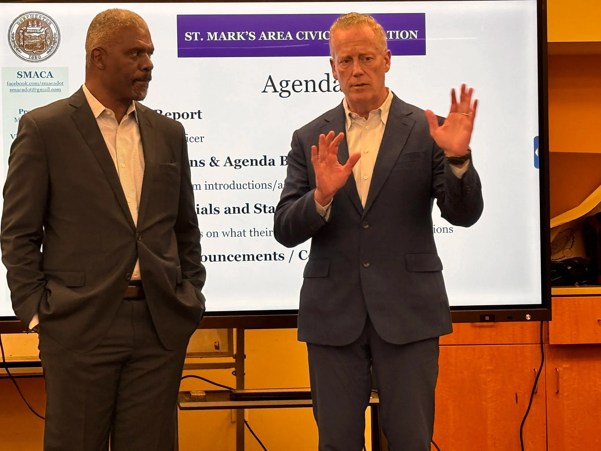 Two men in formal attire are standing in front of a digital screen with a presentation slide about St. Mark's Area Civic Association. One man is speaking while the other is listening.