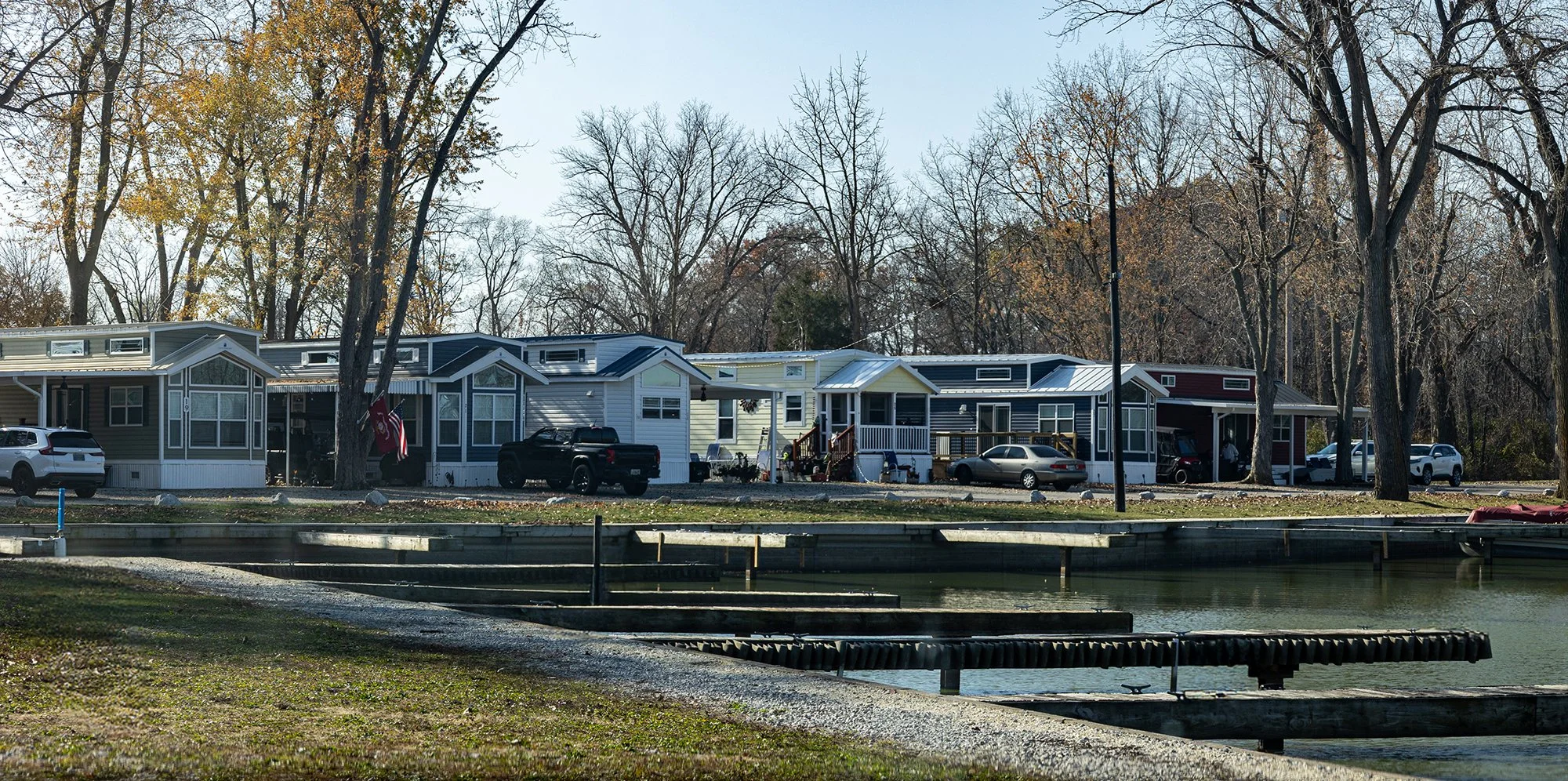 A row of houses near a body of water, with trees in the background and cars parked outside.