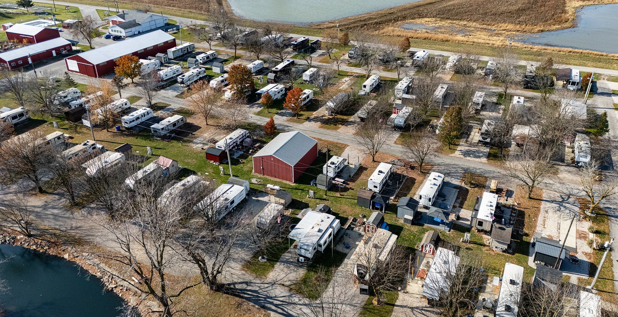 Aerial view of a campground with numerous RVs and trailers parked along roads and in designated spots, surrounded by leafless trees, a pond, and a large red barn building.