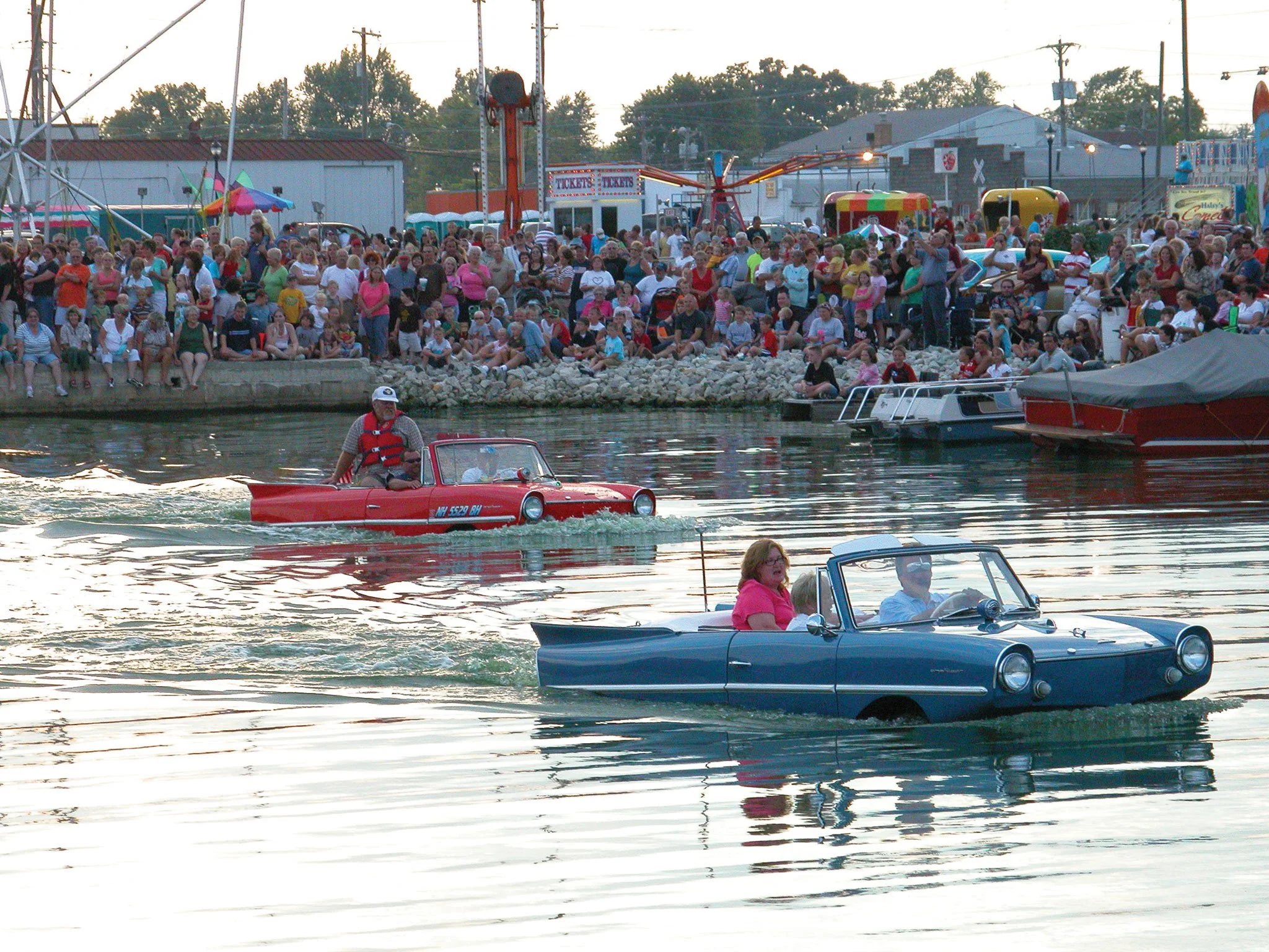 People enjoying a boat parade near a crowded shoreline at an outdoor fair or amusement park.