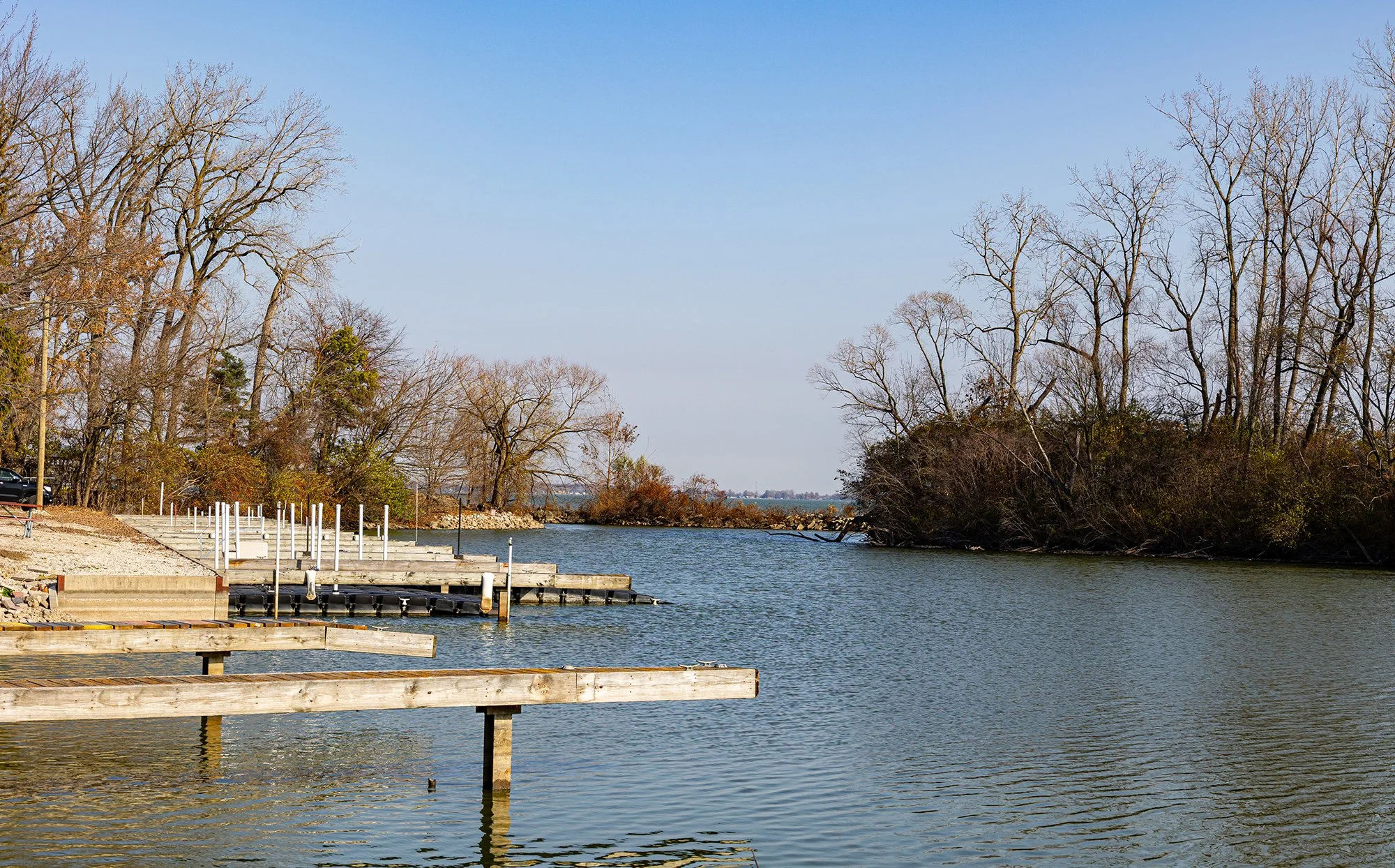 A calm river with wooden docks on the left, leafless trees along the shoreline, and a clear blue sky above.