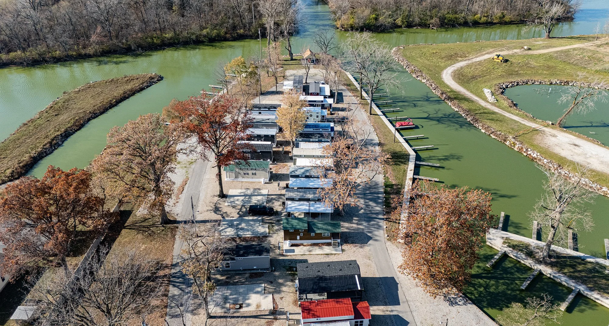 Aerial view of a mobile home park situated along a winding river with some boats, trees with fall foliage, and open grassy areas.