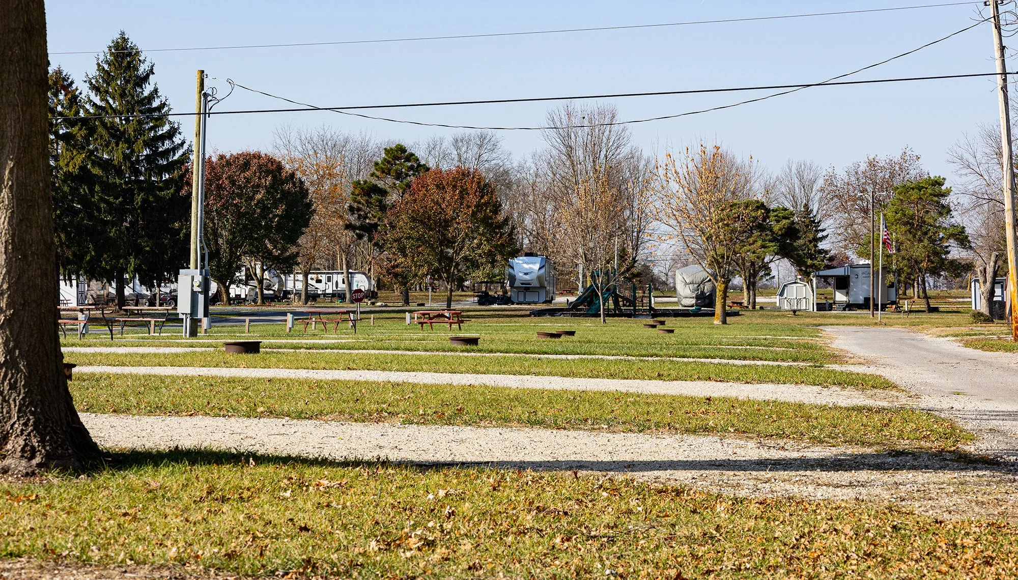 A campground scene with RV campers, trees, picnic tables, and a gravel path, under a clear blue sky.