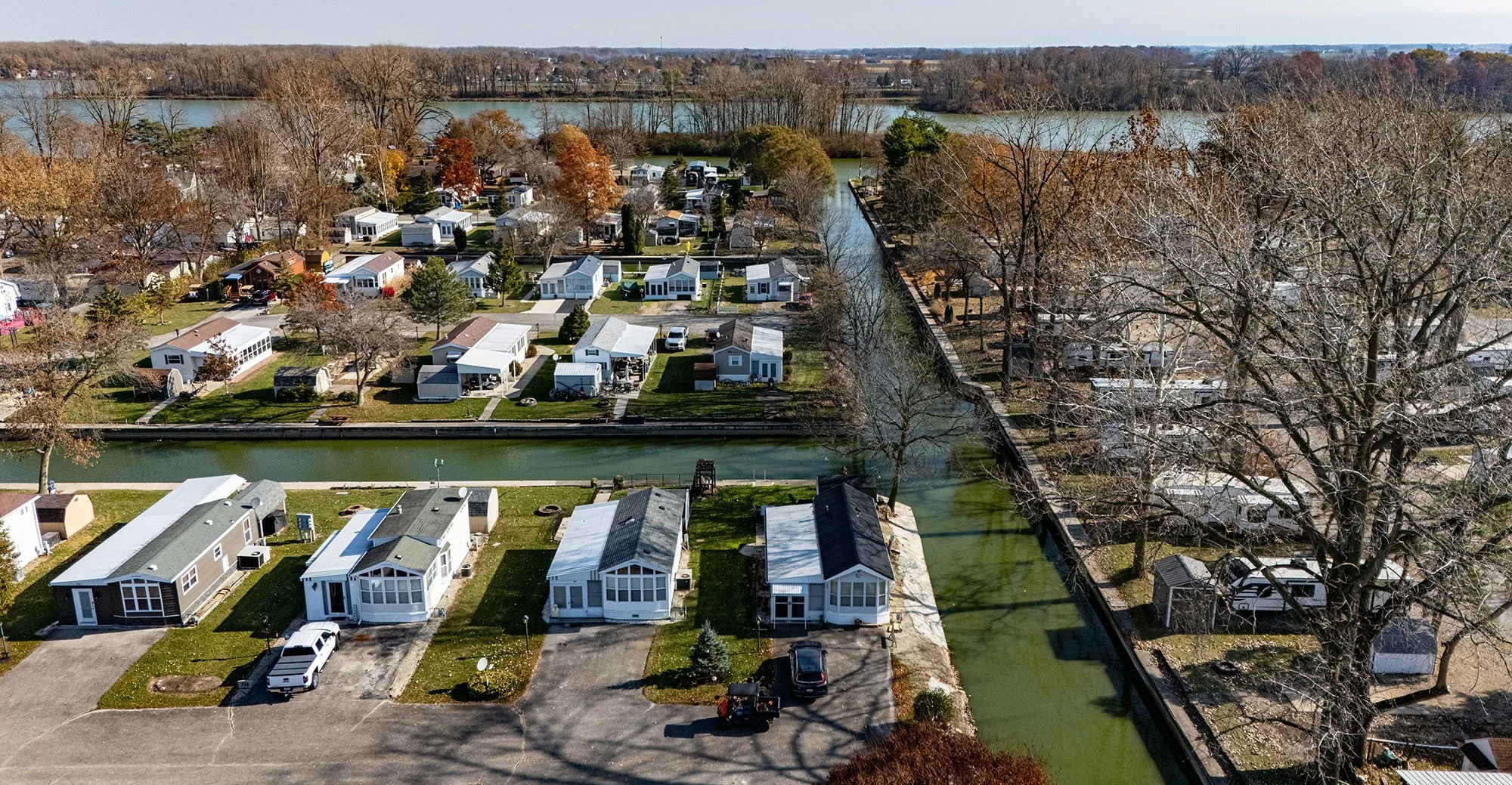 Aerial view of a neighborhood with small houses along a canal, surrounding trees, and a body of water in the background.