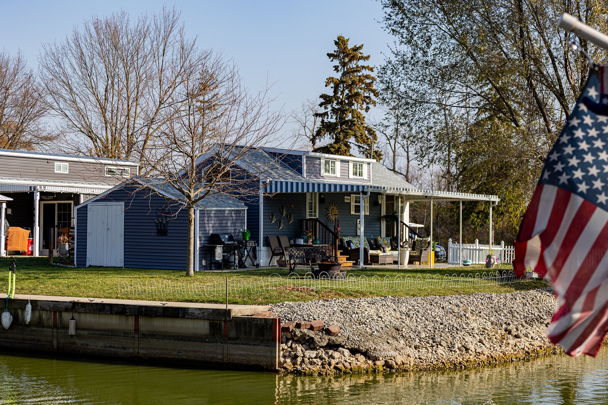 A house with a covered porch facing a body of water, with several trees in the yard, some leafless, and a partial American flag on the right.