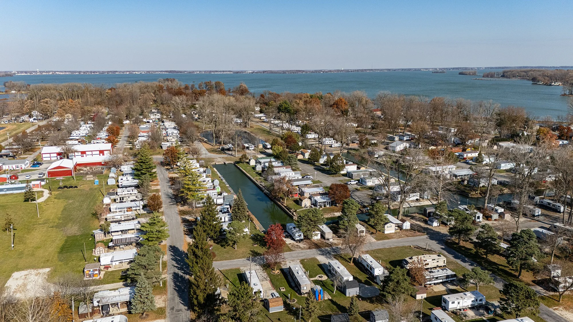 Aerial view of a lakeside RV park with numerous RVs, trees, and water channels, overlooking a large lake or river with a shoreline and trees in the background.