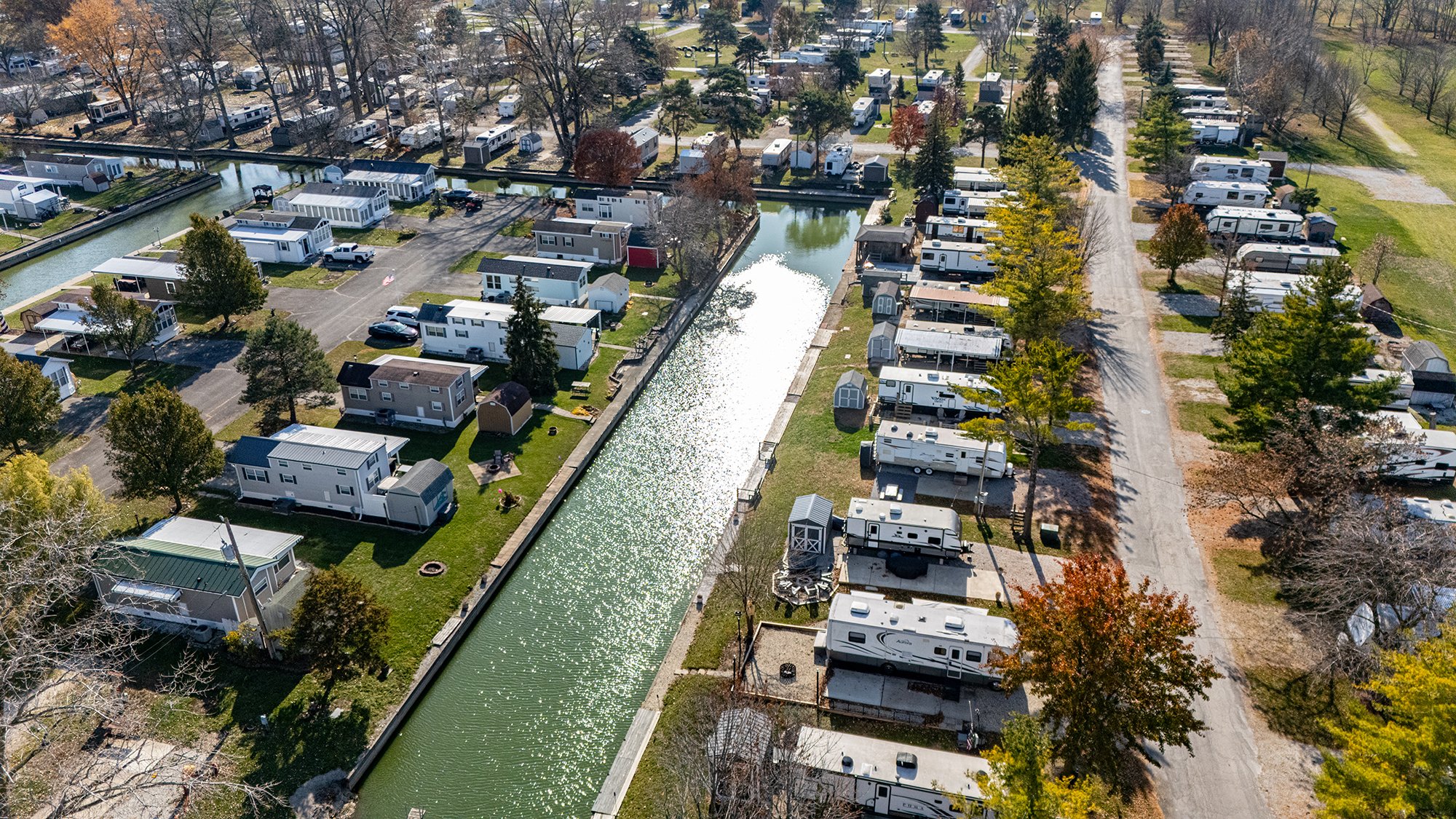 Aerial view of a campground with RVs, trailers, and tiny homes along a canal, surrounded by trees with fall foliage and open grassy areas.