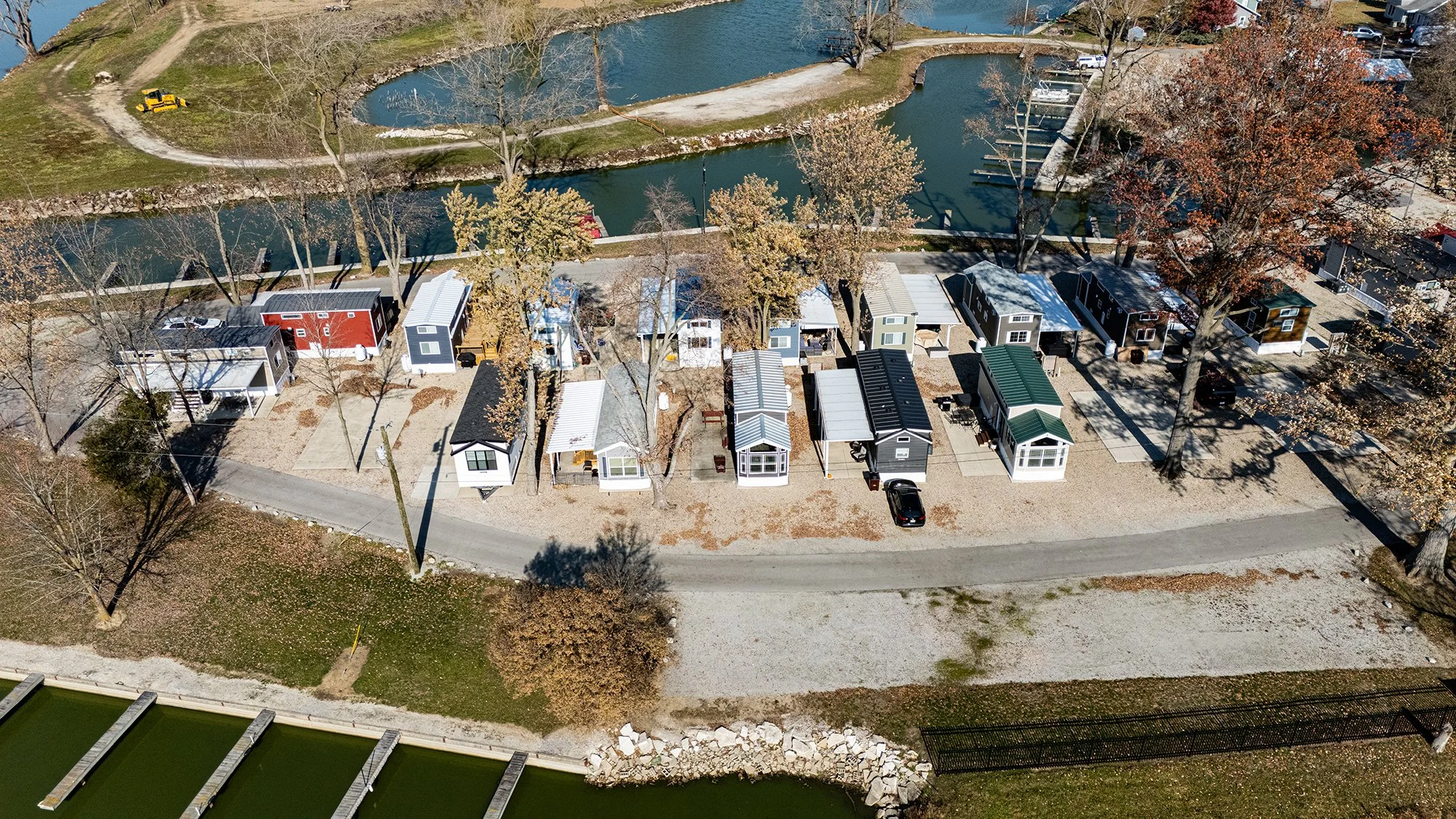 Aerial view of tiny homes along a gravel pathway near a water canal, with trees and parked cars, during autumn.