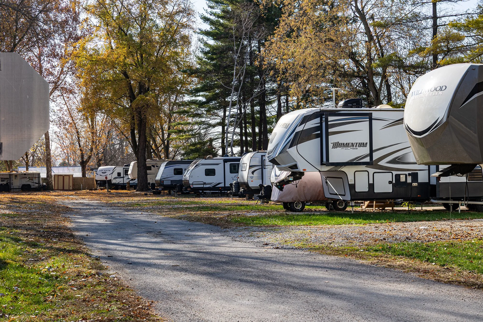 A row of recreational trailers parked on a gravel lot, surrounded by trees with autumn foliage, with some leaves on the ground and a dirt path leading through the scene.