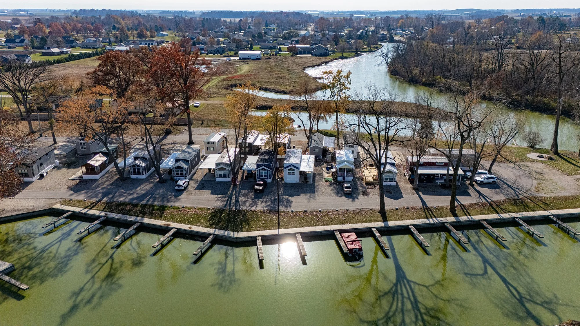 Row of small houses along a waterfront with docks and a boat, leafless trees casting shadows, and a river with a park and houses in the background.
