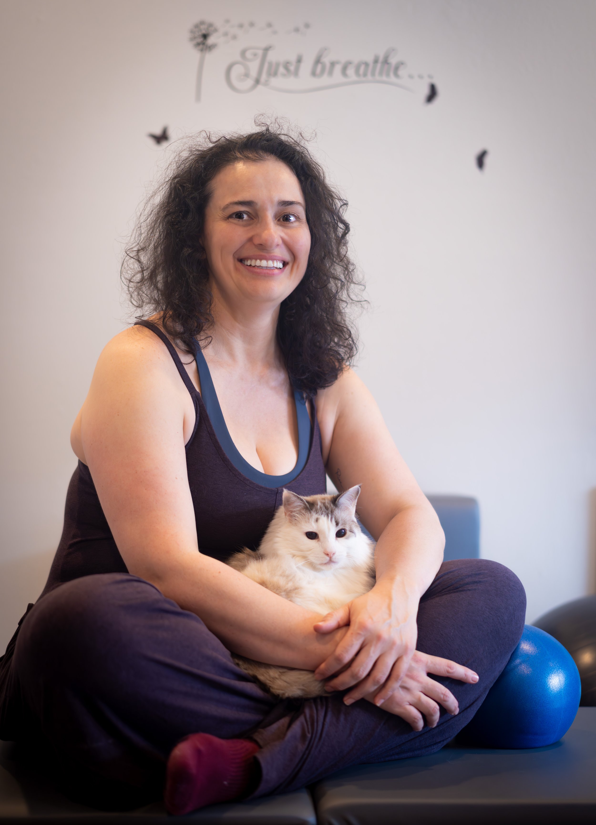 A woman with curly dark hair, smiling, sitting cross-legged on a mat, holding a white and gray fluffy cat on her lap in a fitness or therapy setting, with a wall decor that reads "Just breathe".