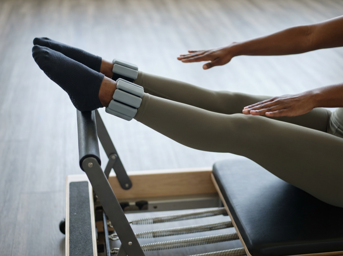 Person exercising on a Pilates reformer machine, with their legs extended and wearing hand and foot resistance equipment.