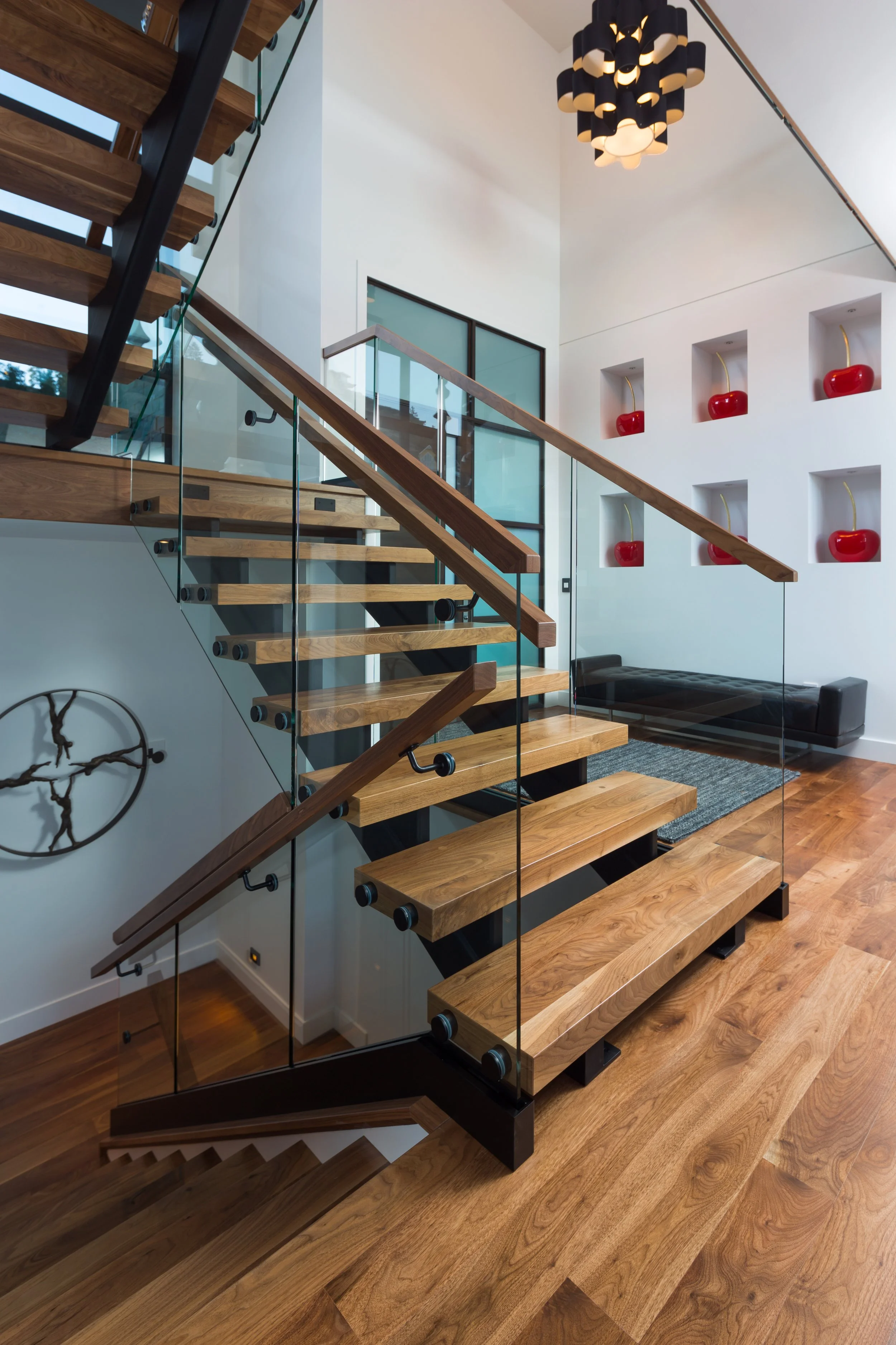 Interior of a modern home featuring a staircase with wooden steps, black supports, and glass railing, a white wall with built-in red decorative objects, a contemporary black and white chandelier, and a black bench on a wooden floor.