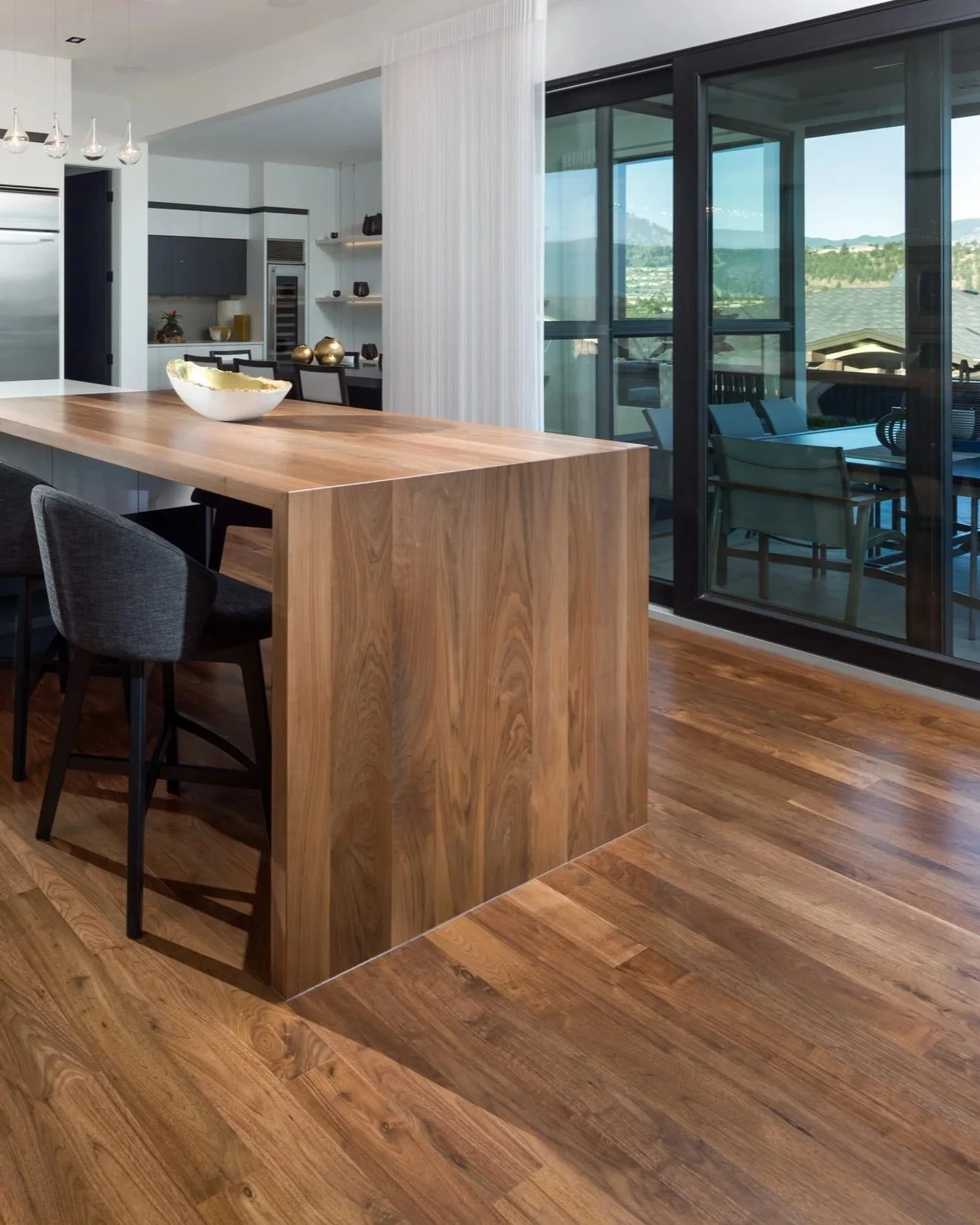 Modern kitchen and dining area with a large wooden island, gray chairs, and a sliding glass door leading to an outdoor patio with mountain views. Beautiful hardwood floors.