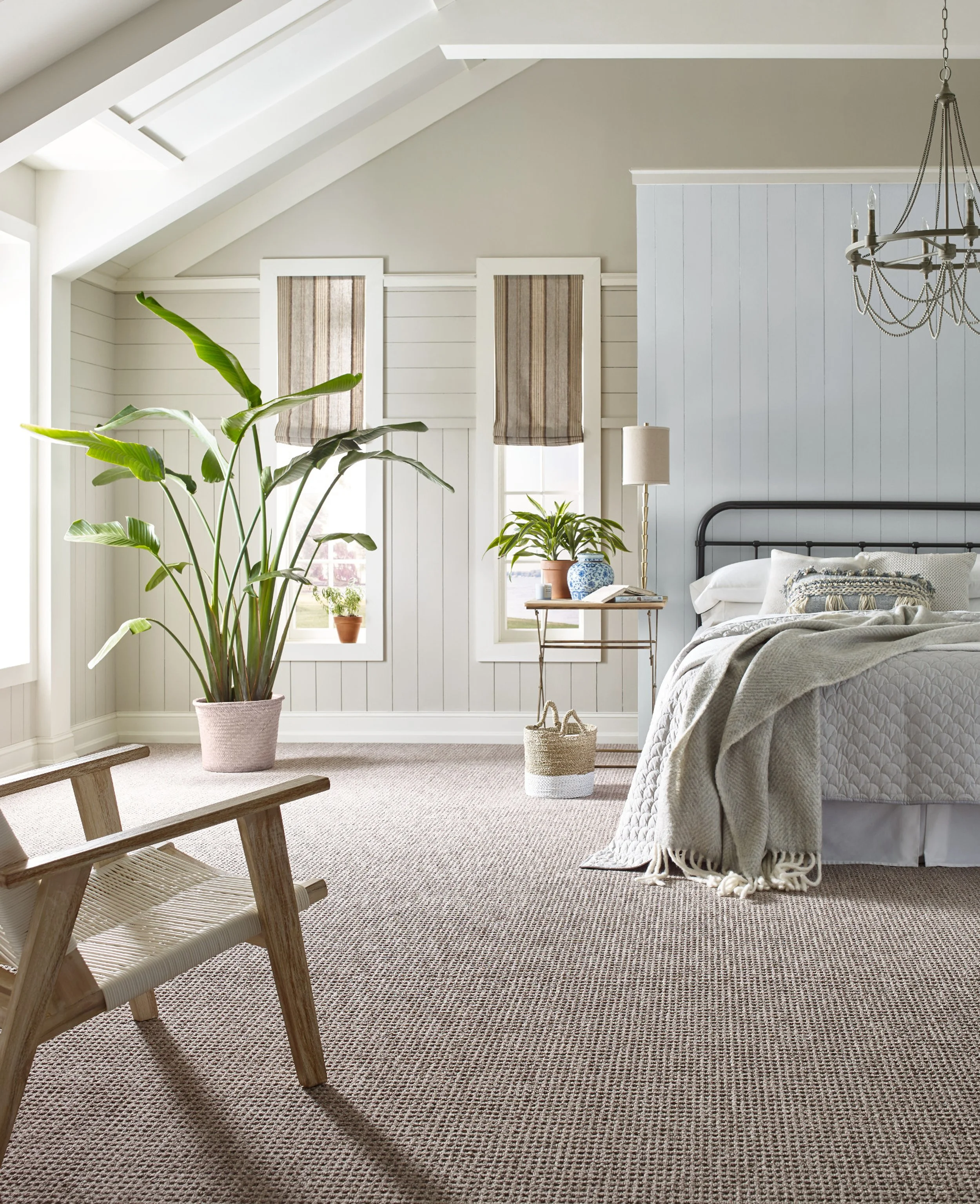 A cozy bedroom with a cozy bed covered in a light blanket and a throw, beside a small side table with a blue-and-white vase, and a large potted plant with green leaves near the window. The room is well-lit with natural light, featuring a textured carpet, wooden bench, and a chandelier.