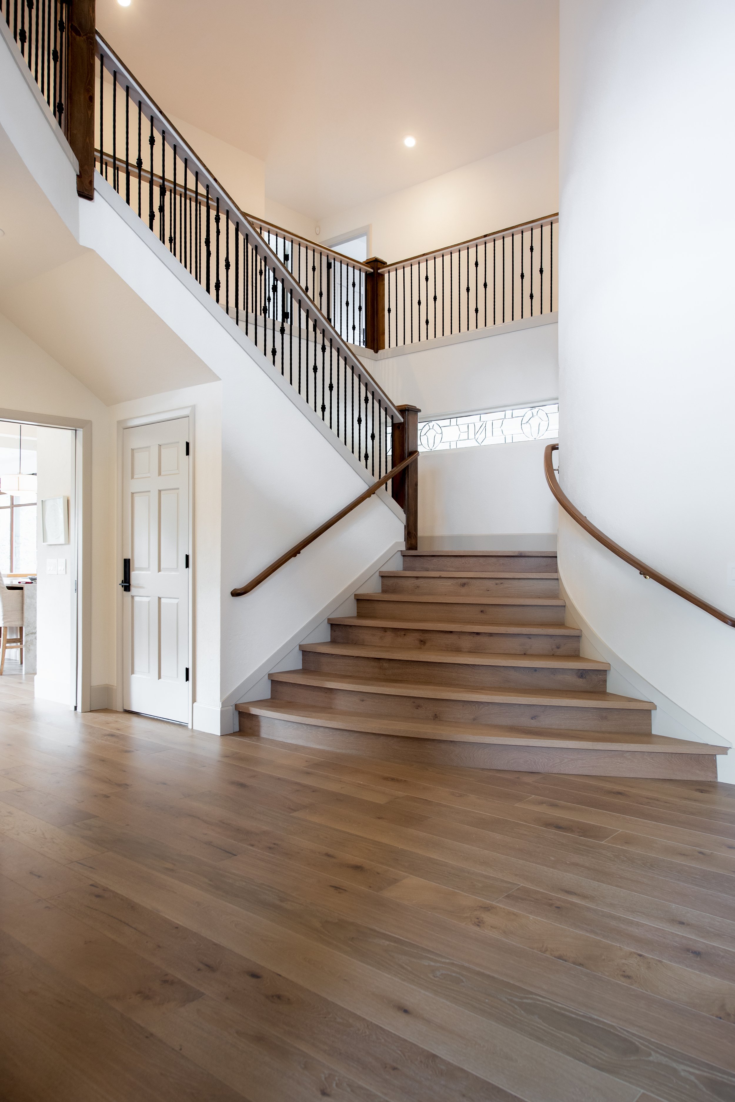 Interior of a house with a staircase featuring wooden steps and black metal balusters, white walls, and a stained glass window near the bottom. Beautiful hardwood stairs.