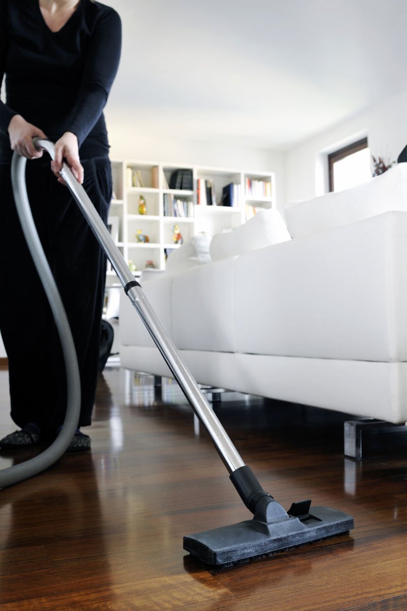 Person vacuuming hardwood floor in living room with white sofa and bookshelf in background.