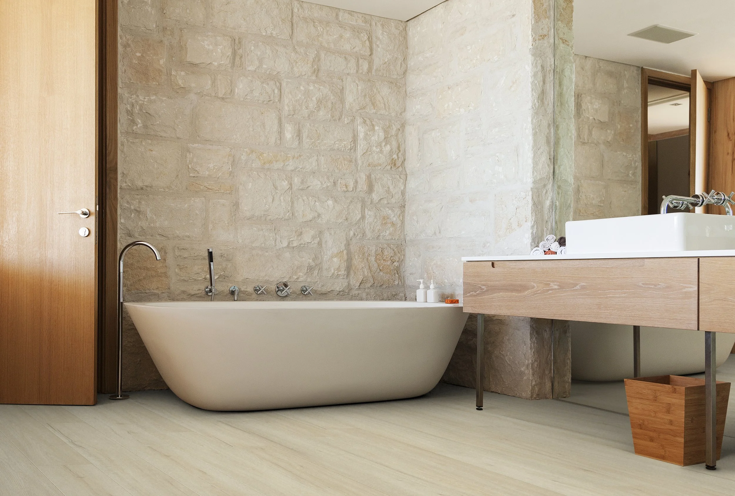 A modern bathroom features a sleek, white bathtub with a chrome faucet and fixtures, set against a textured beige stone wall. To the right, a wooden vanity with a white rectangular sink and chrome two-handle faucet is visible, along with rolled towels and soap dispensers. A large mirror is mounted above the sink, and a wooden door is to the left.