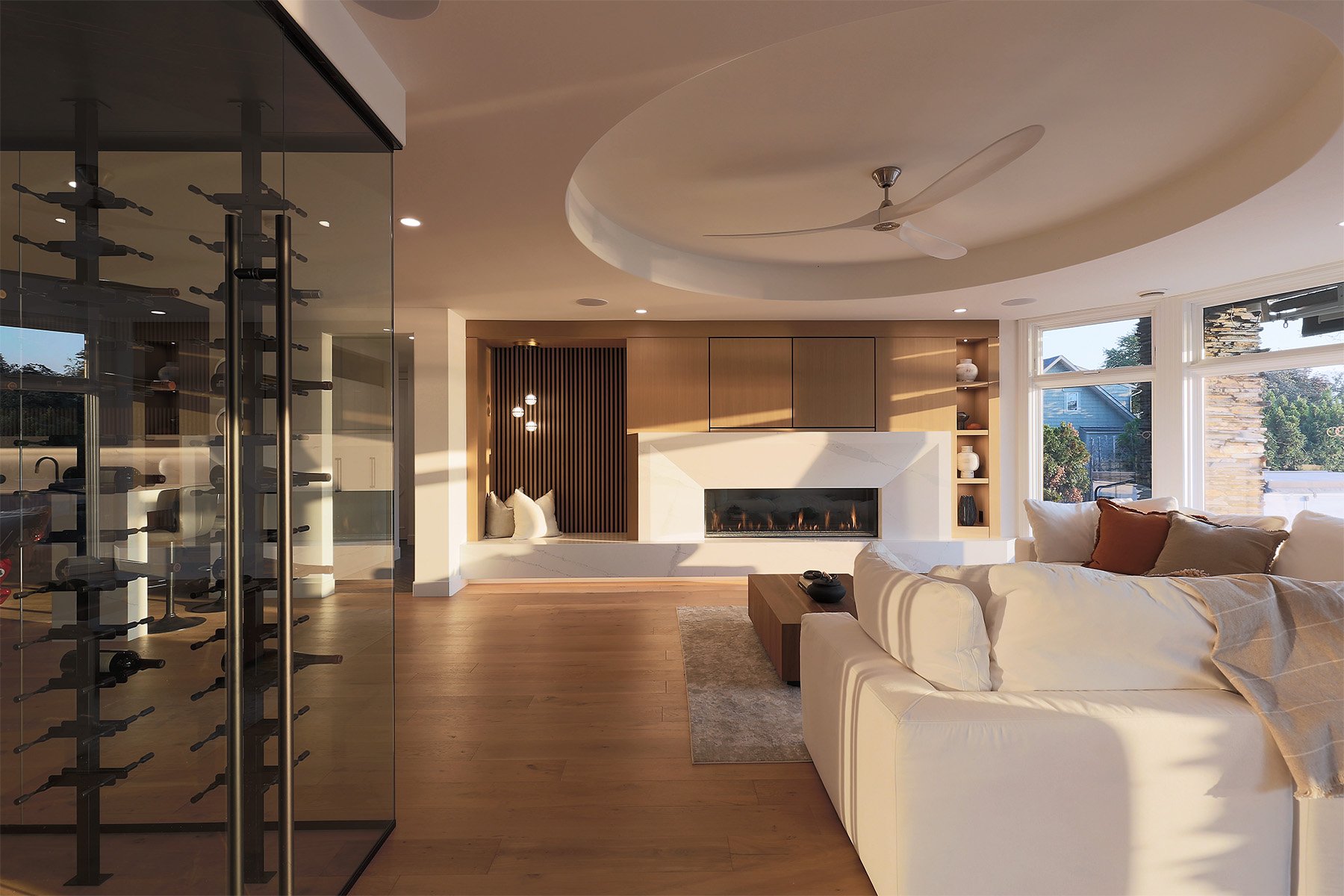 A modern living room with a white sectional sofa, a fireplace, large windows, and a ceiling fan.