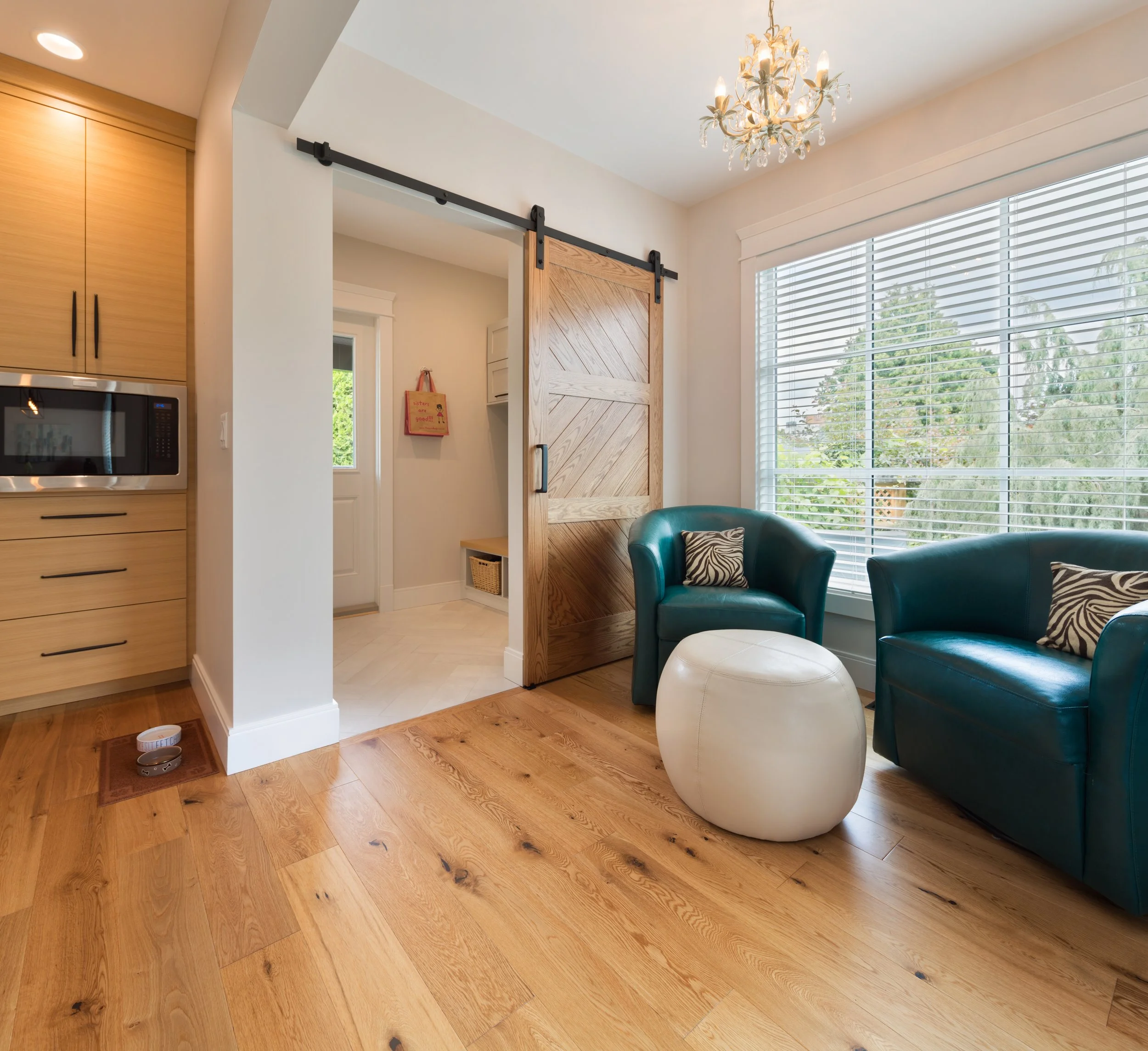 Living room with large window, teal armchairs with zebra print pillows, a white ottoman, a wooden sliding barn door, and a chandelier. Beautiful hardwood floors.