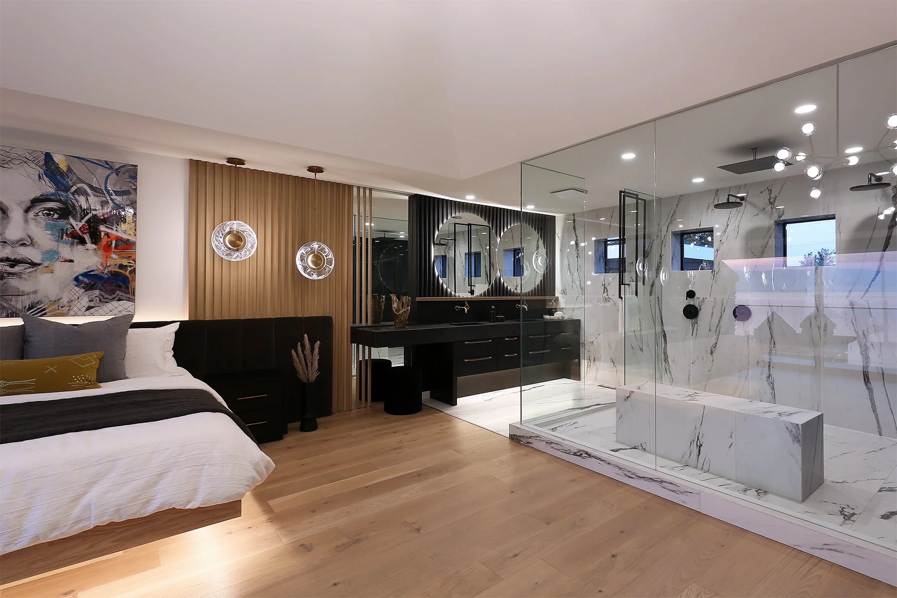 Modern bedroom with a bed featuring white and gray bedding, colorful abstract portrait wall art, wood paneling, circular hanging light fixtures, and a glass-walled bathroom with marble finishes, double sinks, and round mirrors.