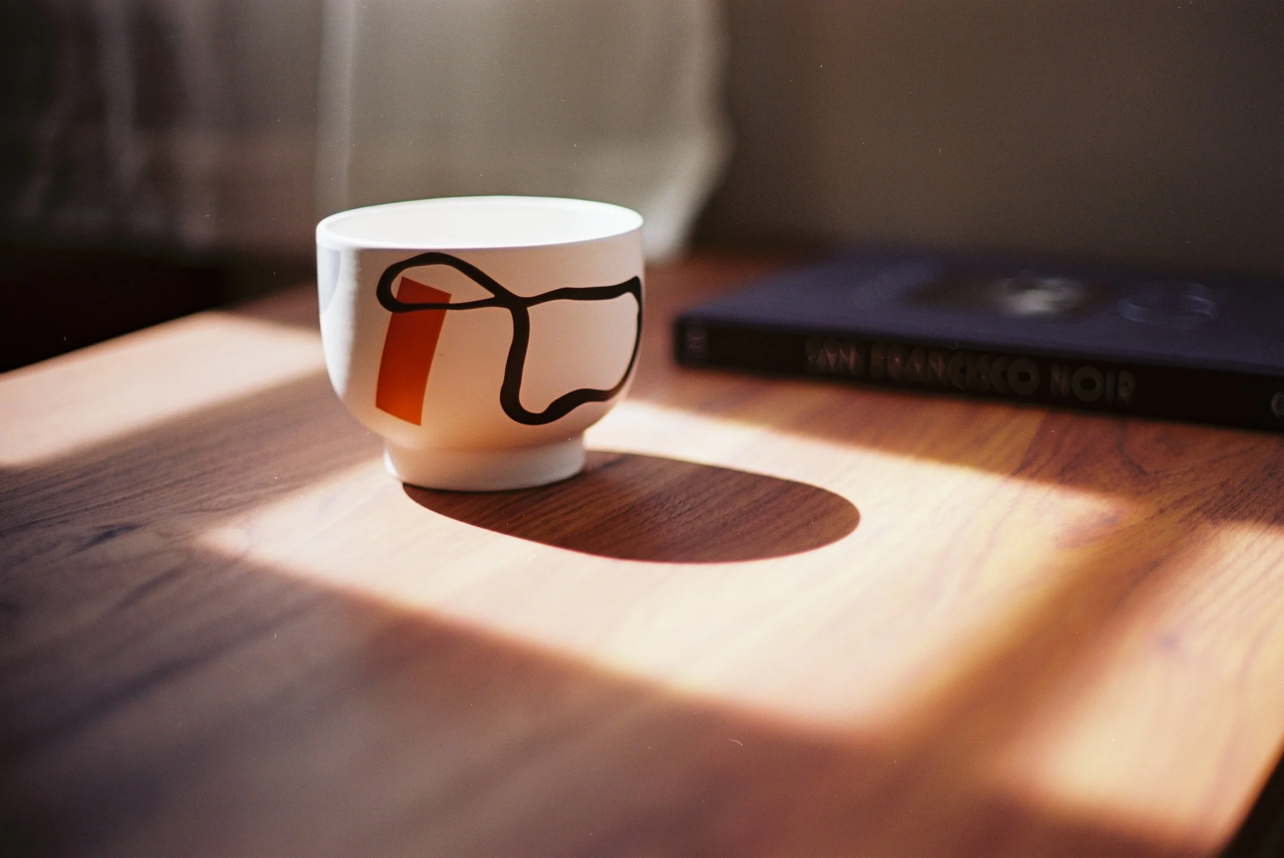 A white ceramic cup with a black outline drawing and the number 1 in orange, placed on a wooden table with sunlight casting a shadow. In the background, there is a closed black book titled 'GIRL, WOMAN, OTHER' lying on the table.