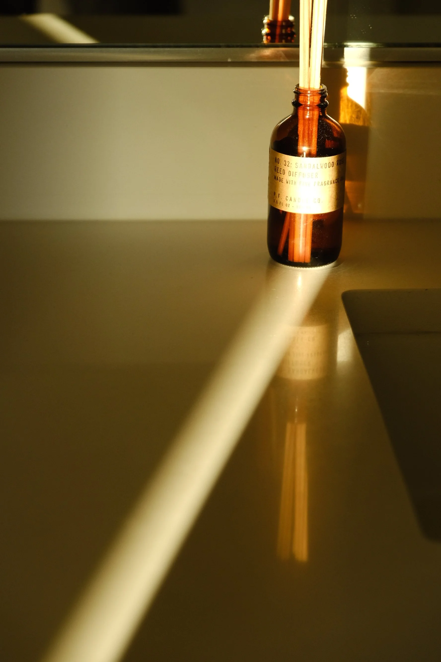 A small amber glass bottle with a gold label and a reed diffuser stick inside, sitting on a reflective surface with a shadow cast behind it, reflected in a mirror.