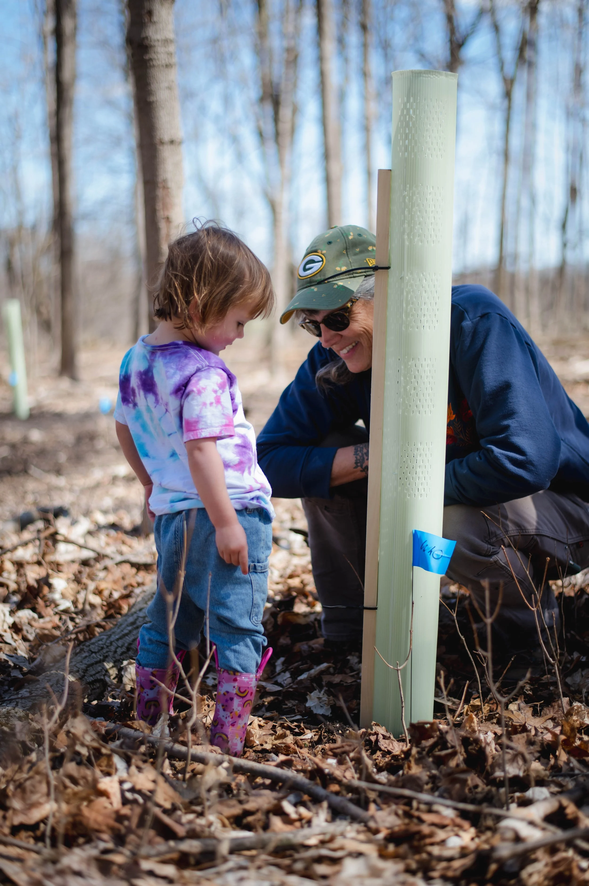 A woman crouches next to a young child, smiling, behind a tree seedling planted in a cage