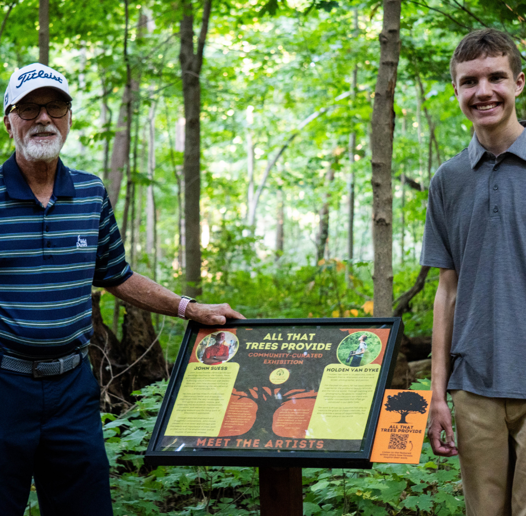 An older man and a teenage boy stand in the Forest Exploration Center on a warm October day surrounding an introductory panel for their art exhibition along the Community Curated Trail Spur