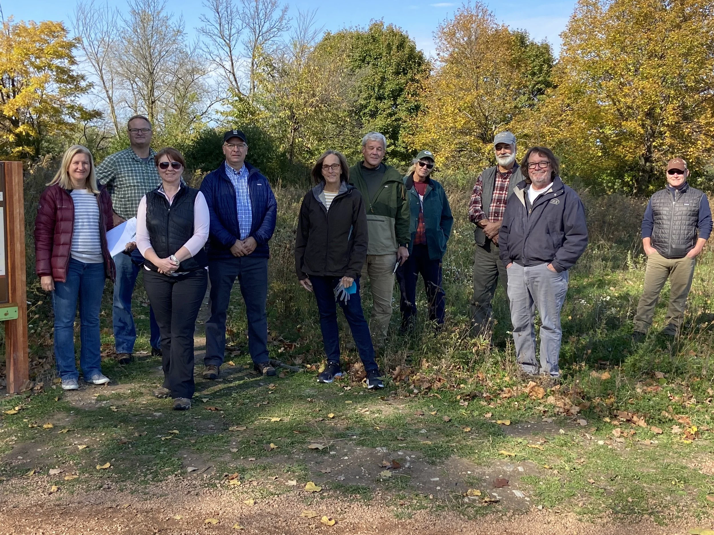 A group of board members, volunteers, and staff from the Forest Exploration Center stand next to the trailhead on a sunny fall day