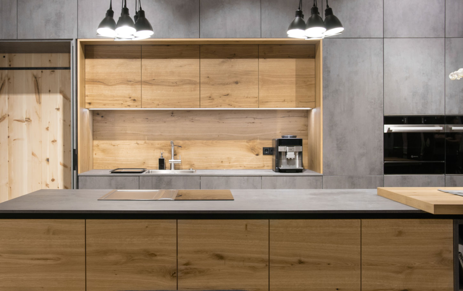 Moderne grifflose Küche mit Holzelementen in Eiche und Fronten in Betonoptik, beleuchtet durch schwarze Deckenlampen, mit Spüle, Kaffeemaschine und einer offenen Ablage, Küchenzeile mit integierter Speistür, große Insel, grifflose Küche