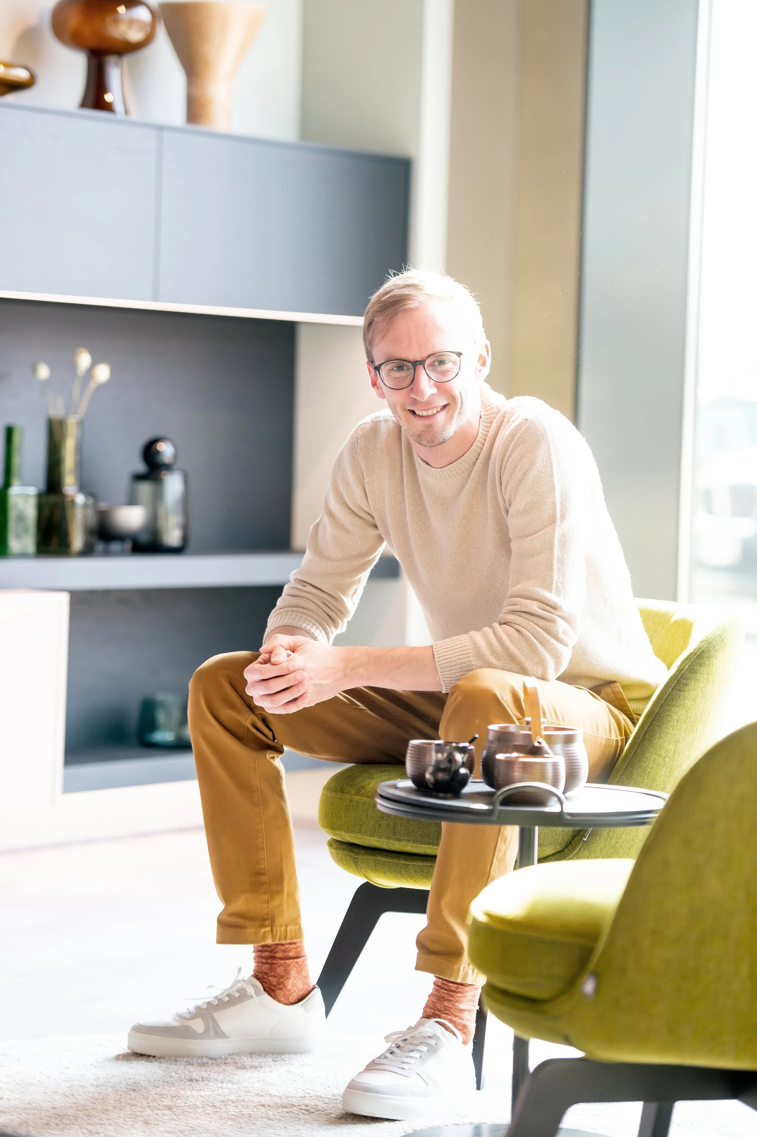 Ein junger Mann mit Brille sitzt in einem modernen Wohnzimmer, trägt einen beigen Pullover und braune Hosen, vor ihm auf einem kleinen Tisch befinden sich Tassen und eine Kaffeemaschine.