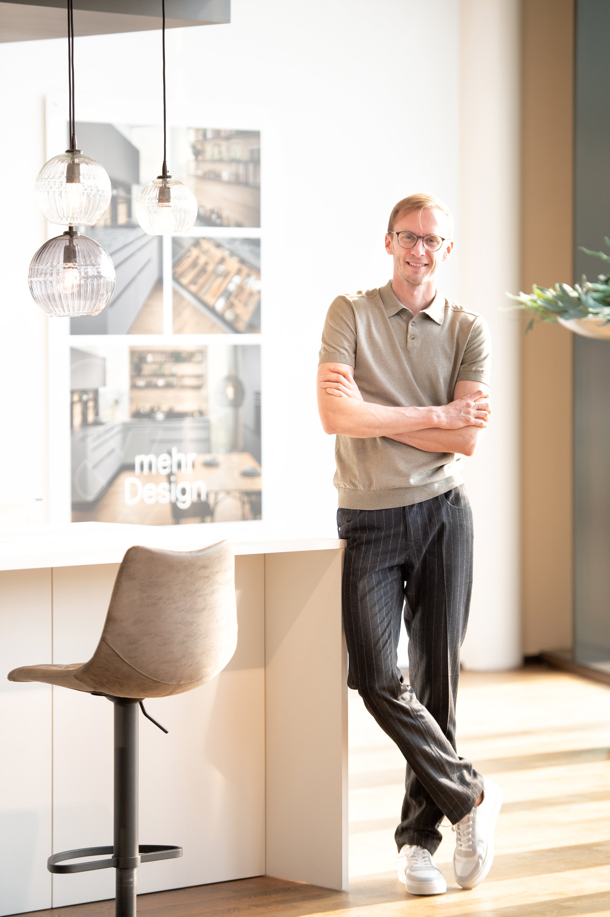 Ein Mann mit Brille, hellbraunen Haaren und lockigem Blick, trägt ein beiges Polo-Shirt und schwarze gestreifte Hosen, steht in einem modernen, hellen Raum mit natürlichen Lichtverhältnissen, neben einem weißen Schreibtisch und einer Sitzgelegenheit.