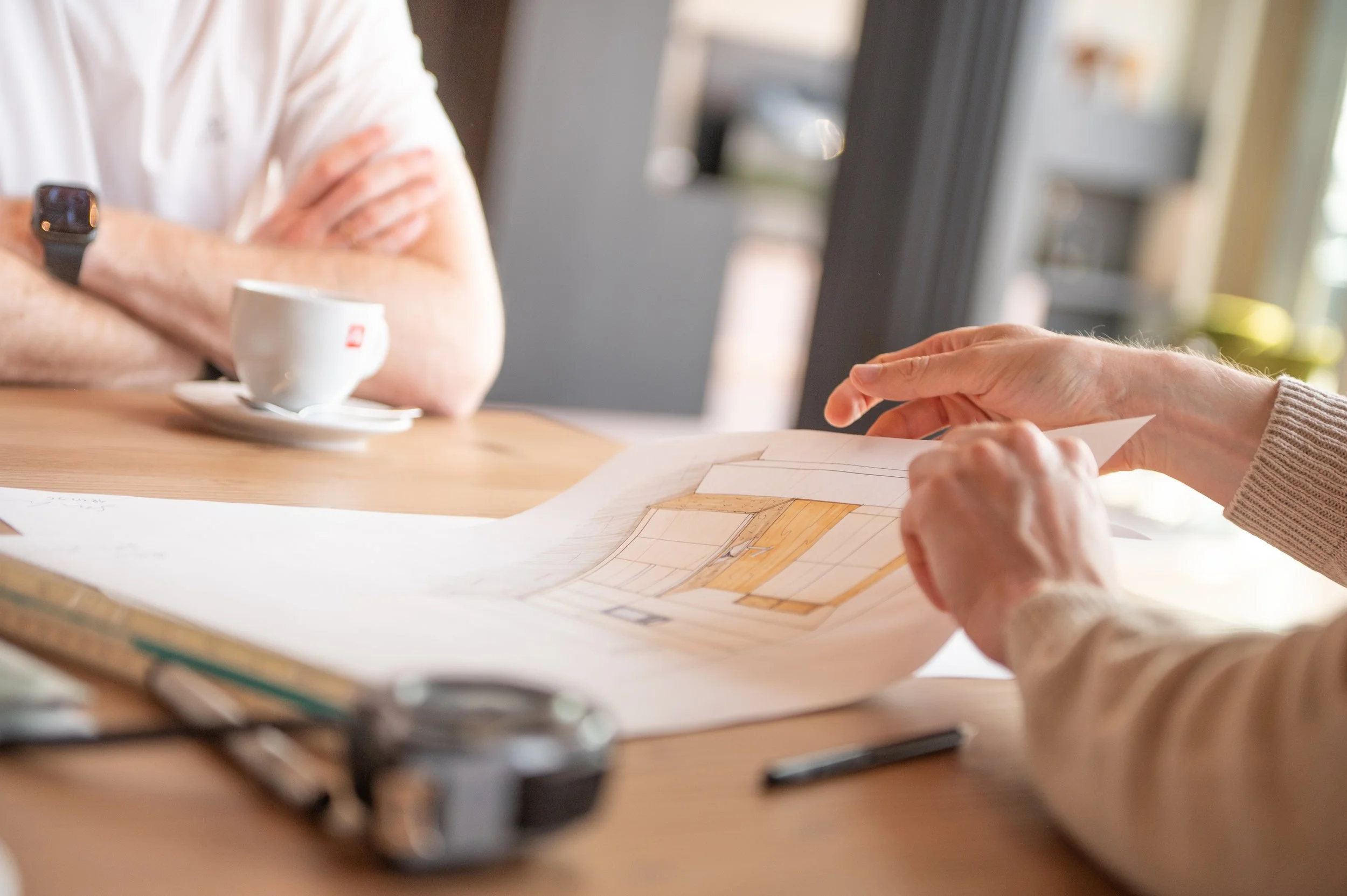 Eine Person zeigt auf einen Entwurf oder Plan, während eine andere Person neben einem Tisch mit einer Tasse Kaffee sitzt. Der Fokus liegt auf den Händen und dem Papier, während eine Person mit verschränkten Armen im Hintergrund sichtbar ist.