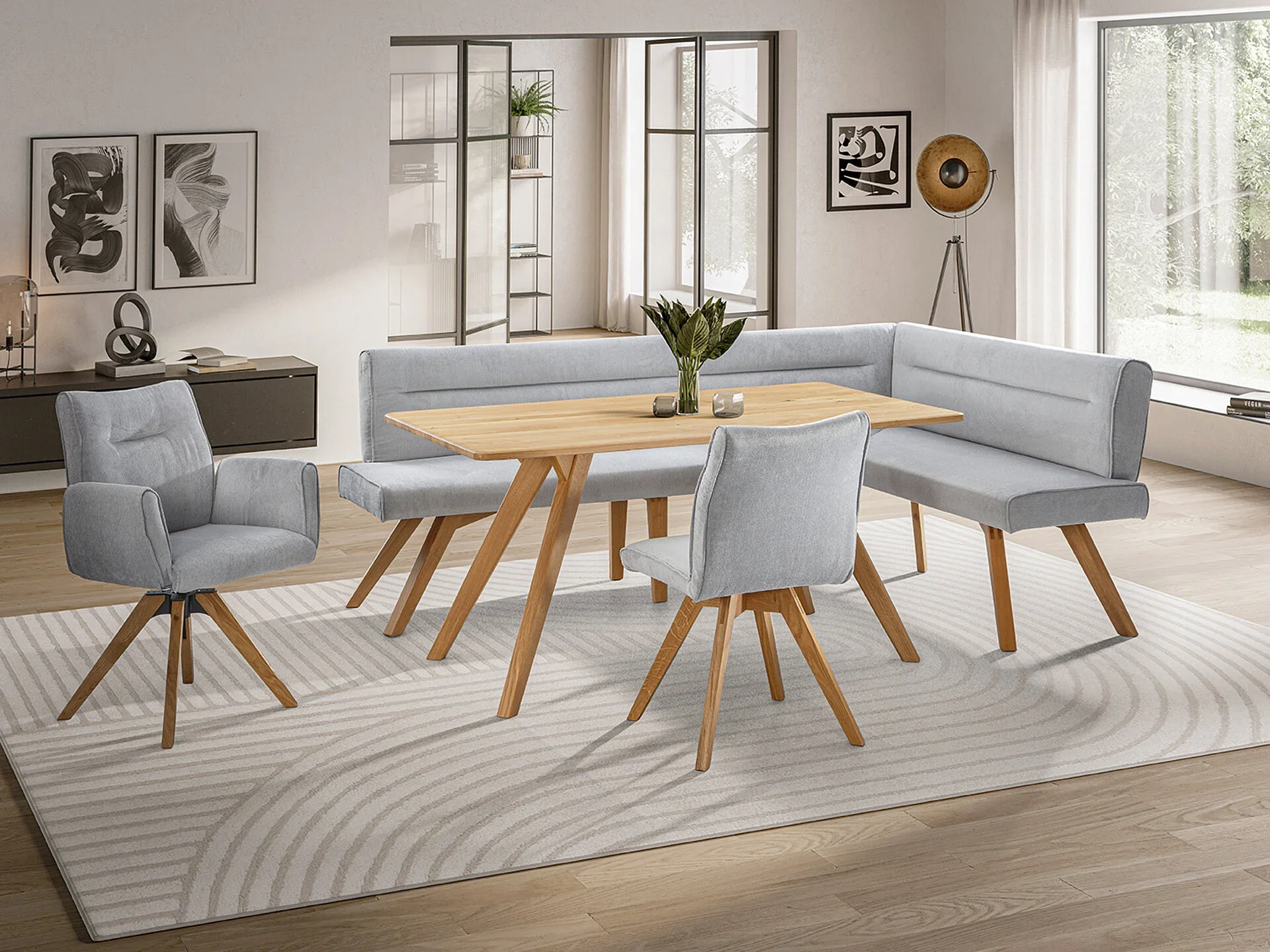 Ein moderner Wohnraum mit einem Esstisch aus Eichenholz, vier grauen Stühlen, einer grauen Eckbank, einem schwarzen Lowboard, Wandkunst und einer Stehlampe auf einem beigefarbenen Teppich auf Holzboden.