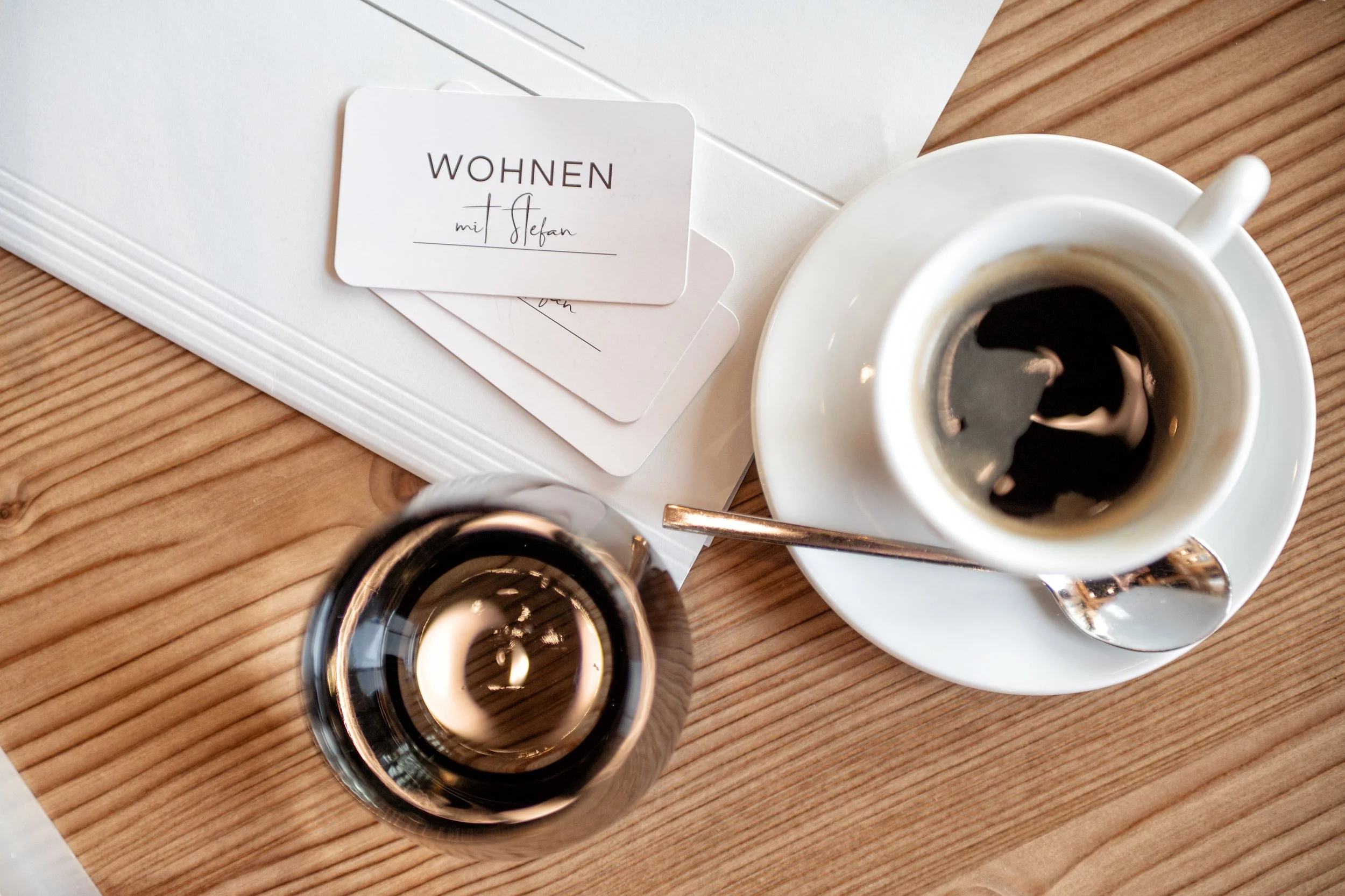 Zwei Tassen schwarzen Kaffee, ein Blatts Papier, eine kleine Schale mit Zucker, ein Löffel und ein Notizbuch auf einem Holz Tisch. Sowie eine Visitenkarte von "Wohnen mit Stefan"
