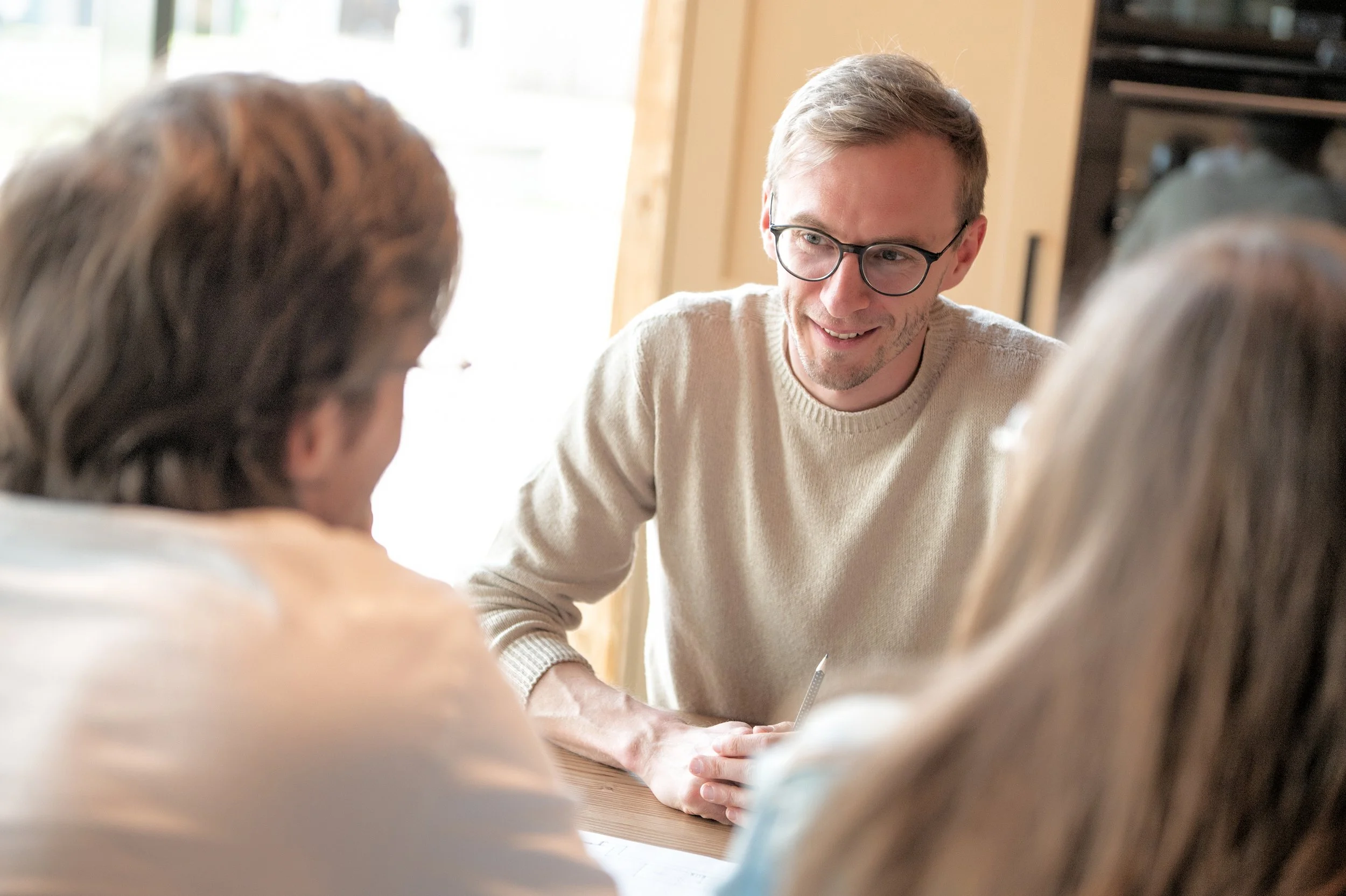 Ein Mann mit Brille und hellbraunem Pullover spricht mit einer Personengruppe in einem Raum mit natürlichem Licht. Beratungsgespräch mit einem jungen Paar bezüglich einer neuen Einrichtung.