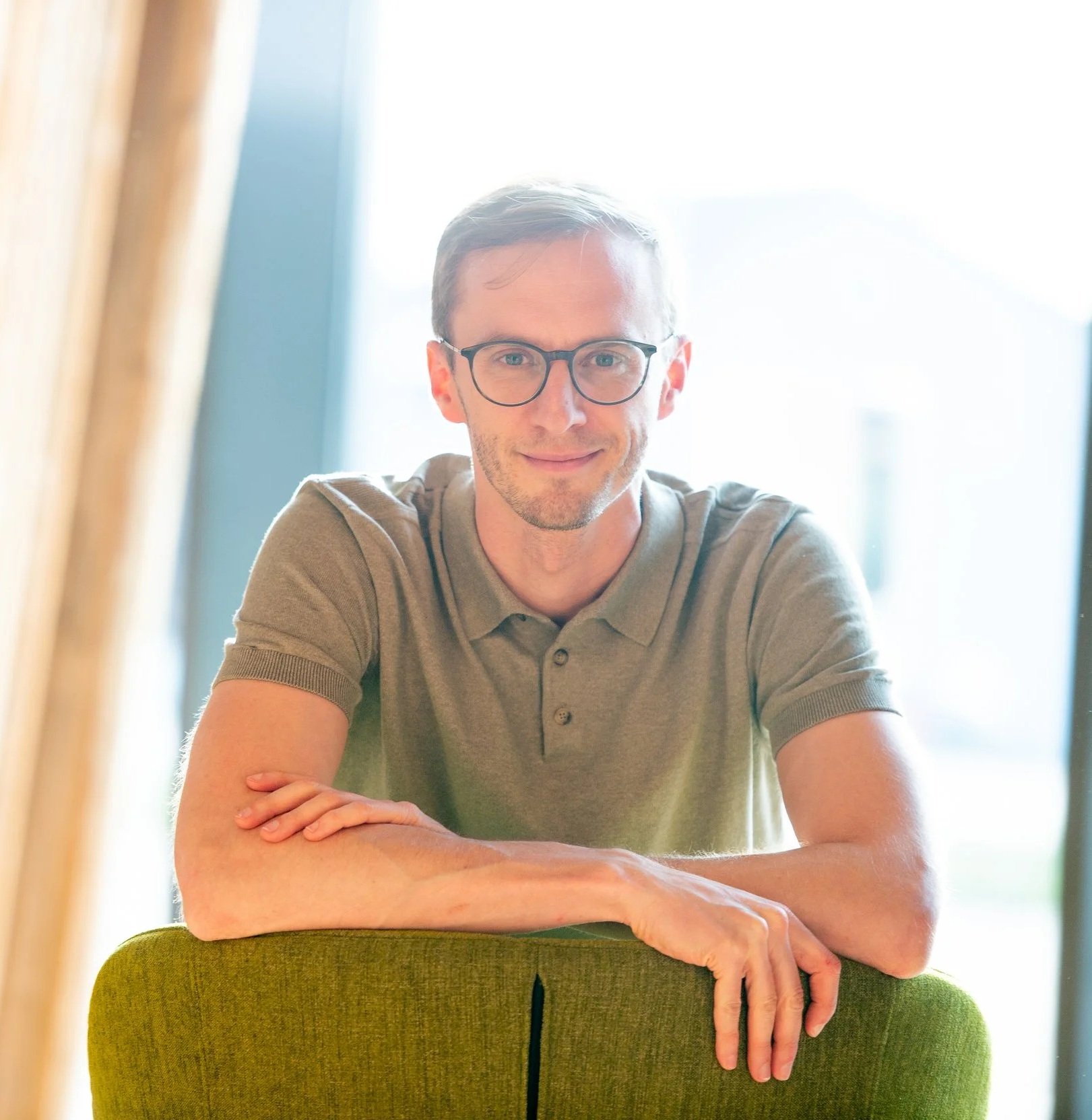 Ein junger Mann mit Brille sitzt hinter einem grünen Hocker und schaut in die Kamera, vor einem hell erleuchteten Hintergrund.