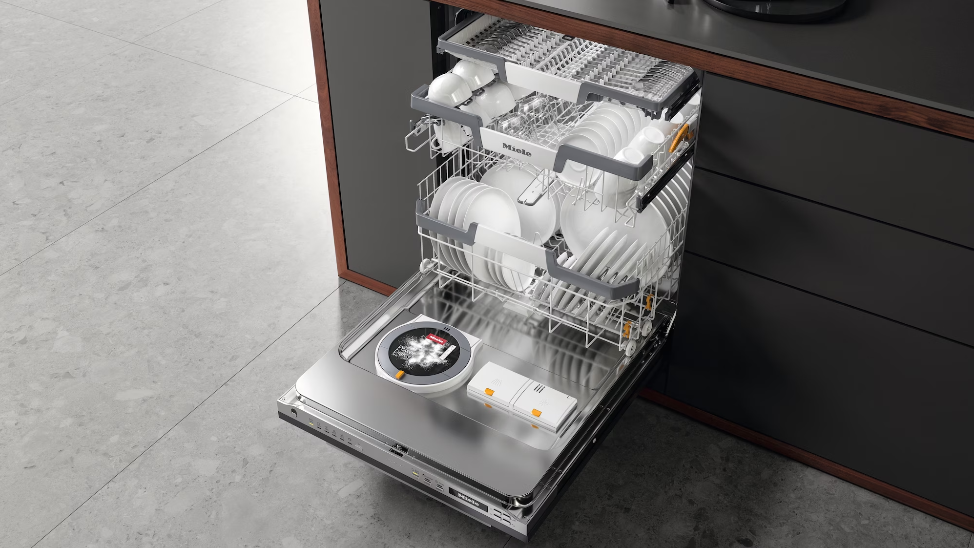 Ein offener Geschirrspüler mit Geschirr und Besteck, eingebaut in eine moderne Küchenschrankfront, neben einem grauen Boden. Miele Geschirrspüler mit AutoDos und Bestecklade