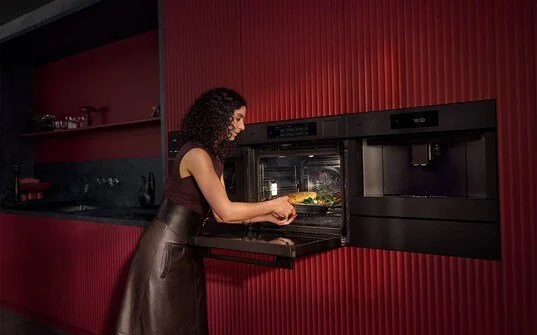 Frau backt im modernen Küchenofen in einer roten Küche. AEG Multidampfgarer mit Wärmelade, sowie AEG Einbaukaffeemaschine, Lamellenfronten