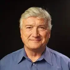 Portrait of an older man with gray hair wearing a blue collared shirt, smiling slightly against a dark background.