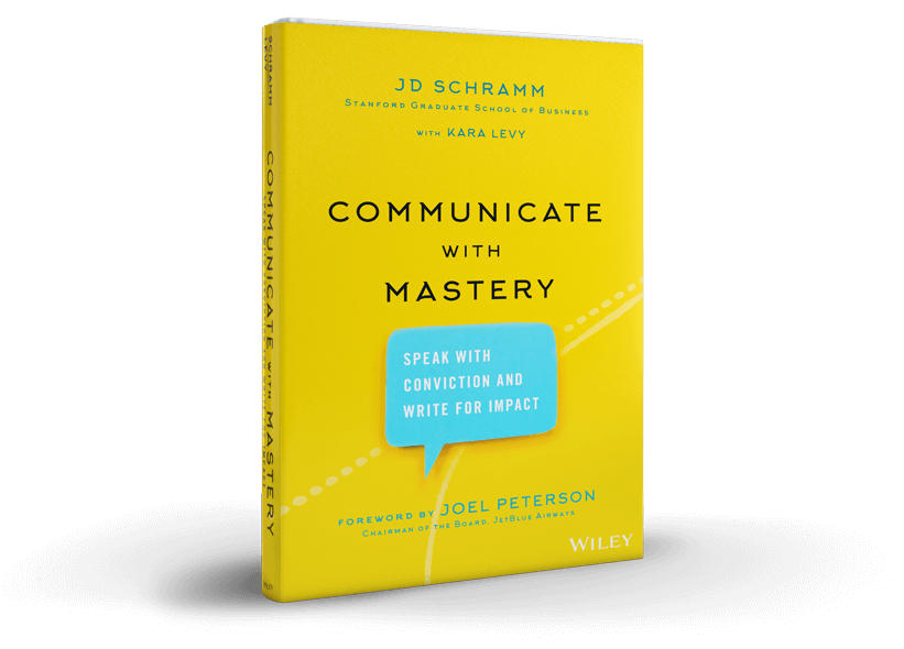 Yellow book titled 'Communicate with Mastery' by JD Schramm with Kara Levy, featuring a blue speech bubble with the text 'Speak with conviction and write for impact' on the cover.